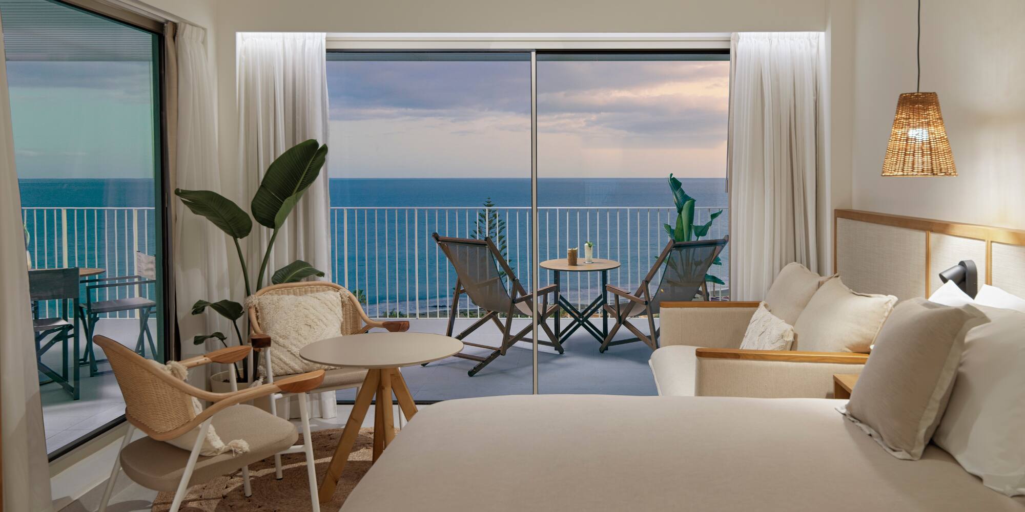 a room with a bed and chairs and a table with chairs and a balcony overlooking the ocean