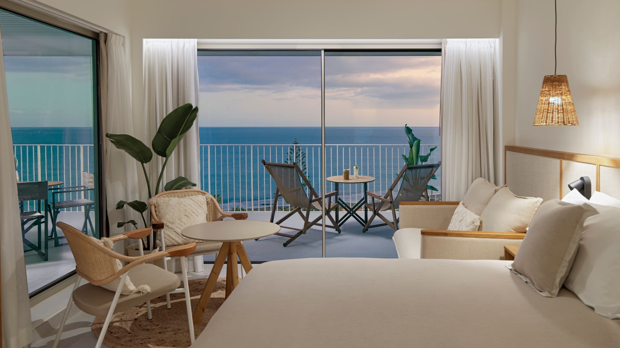 a room with a bed and chairs and a table with chairs and a balcony overlooking the ocean