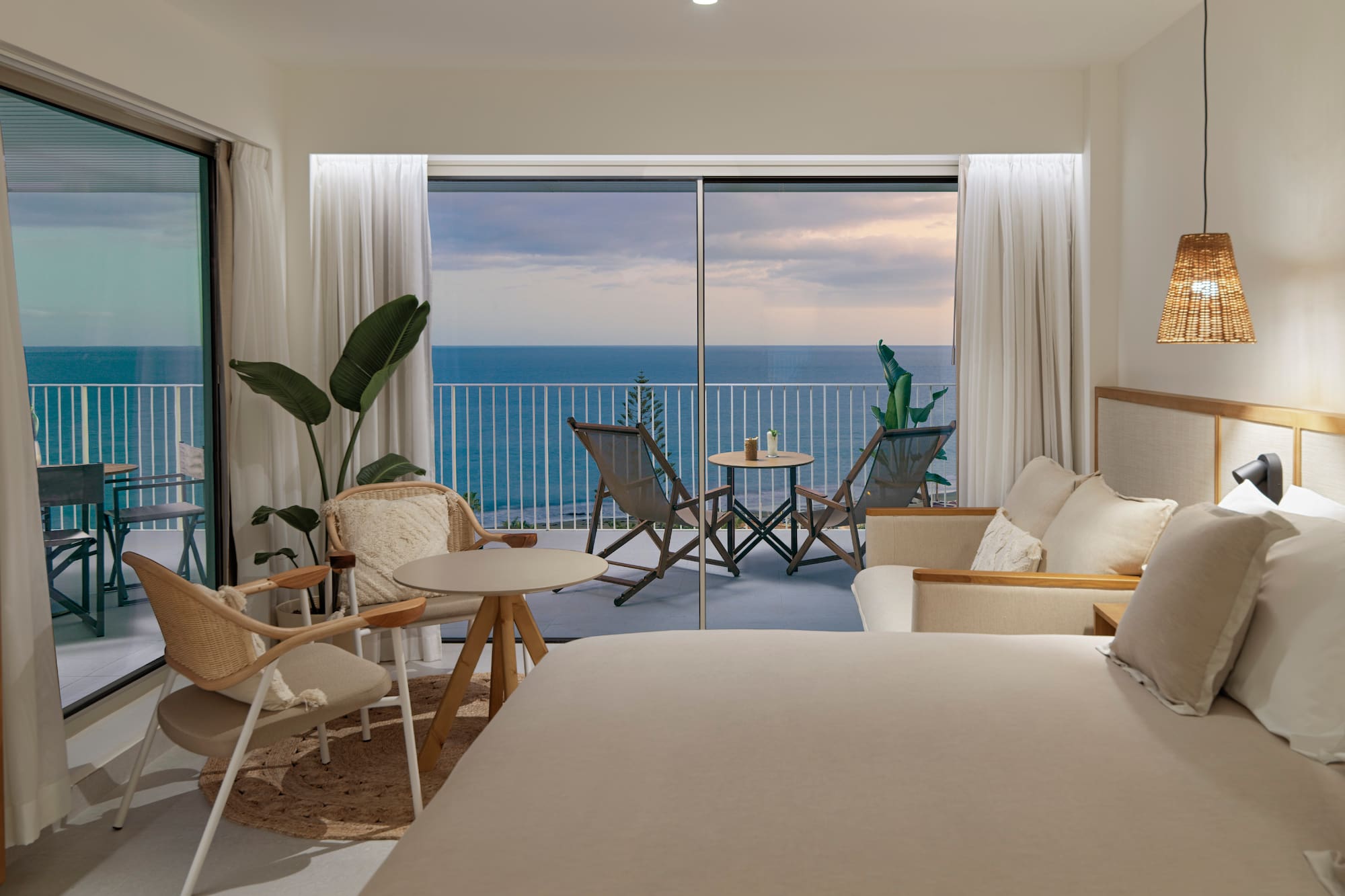 a room with a bed and chairs and a table with chairs and a balcony overlooking the ocean