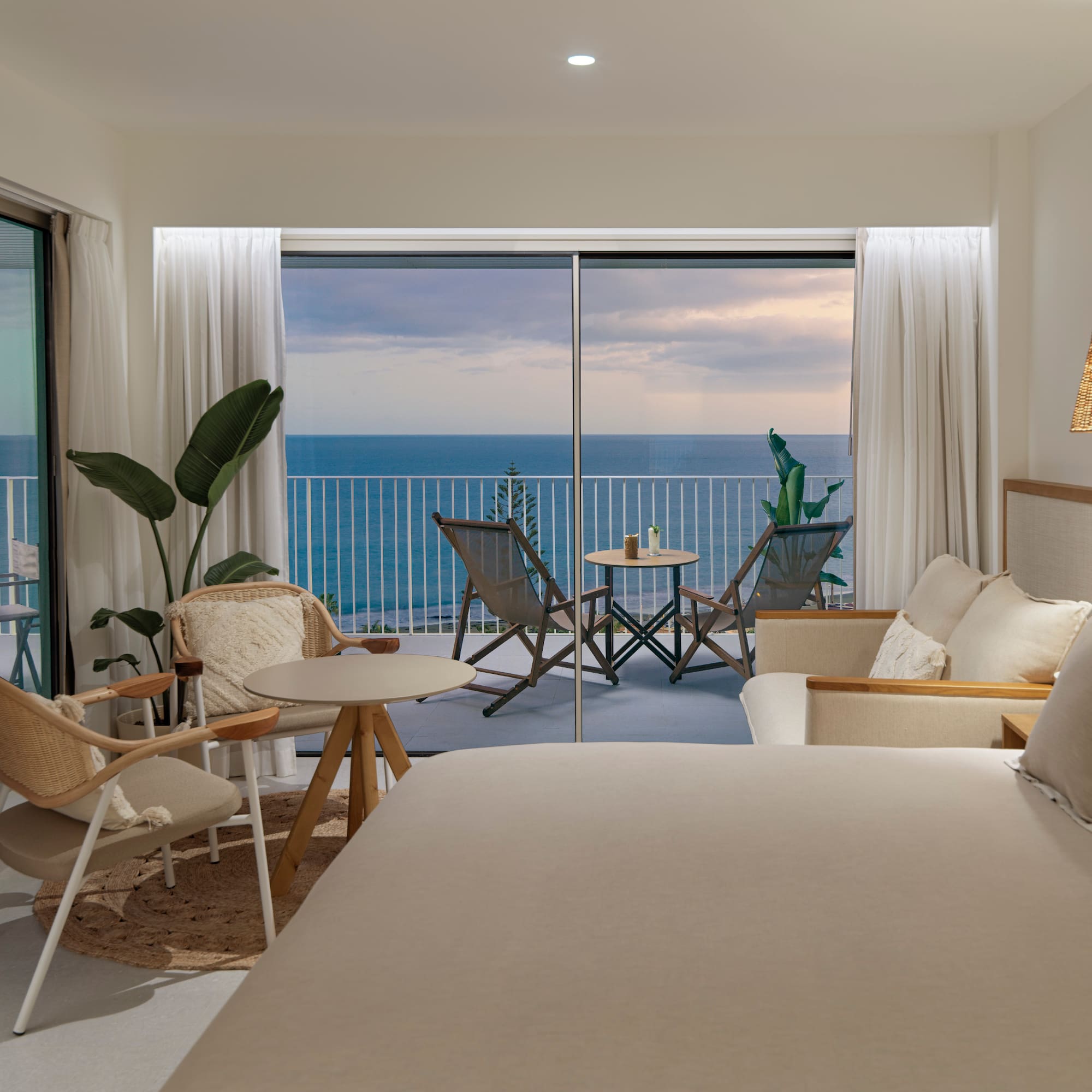 a room with a bed and chairs and a table with chairs and a balcony overlooking the ocean