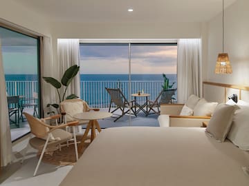 a room with a bed and chairs and a table with chairs and a balcony overlooking the ocean