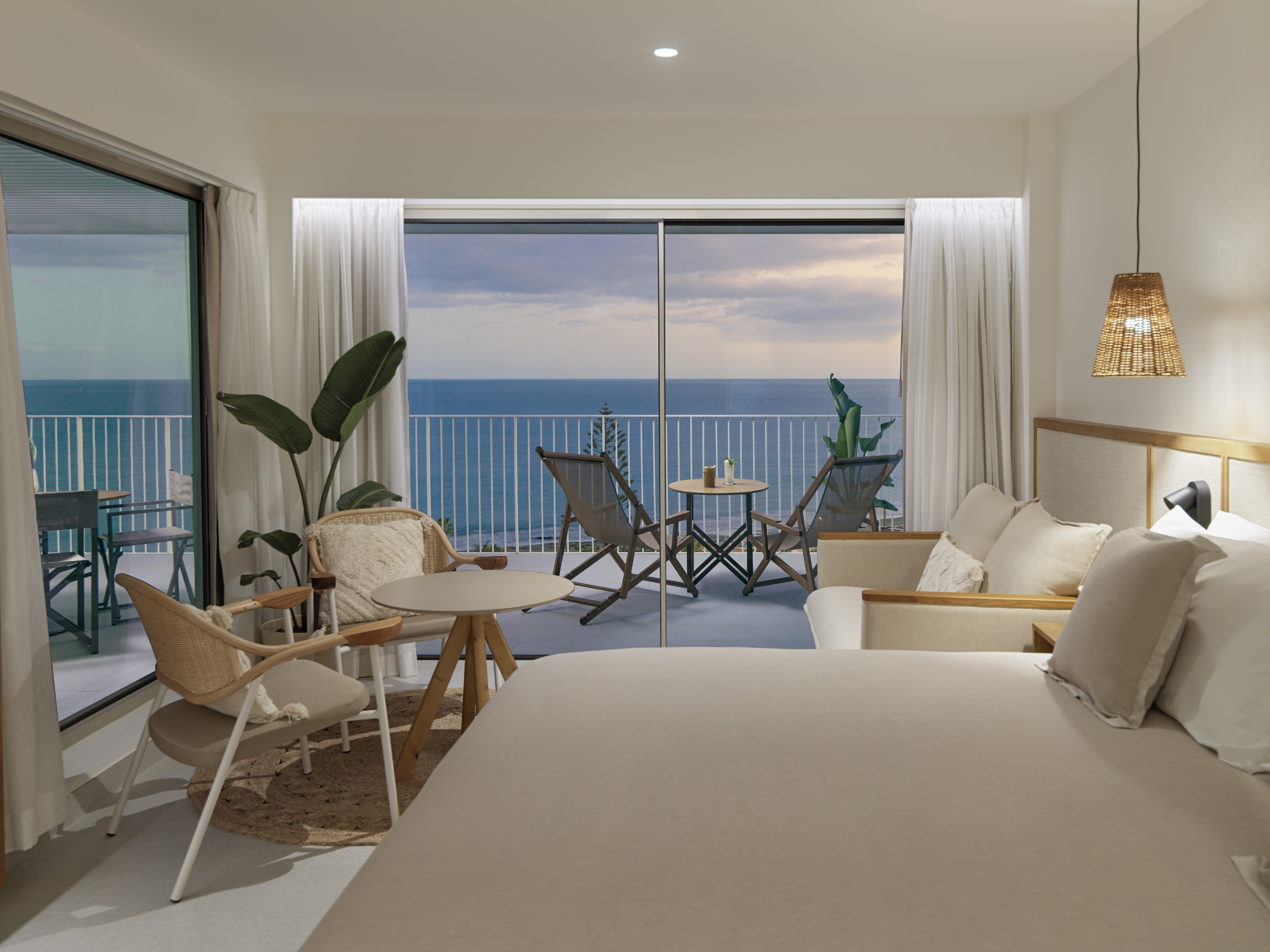 a room with a bed and chairs and a table with chairs and a balcony overlooking the ocean