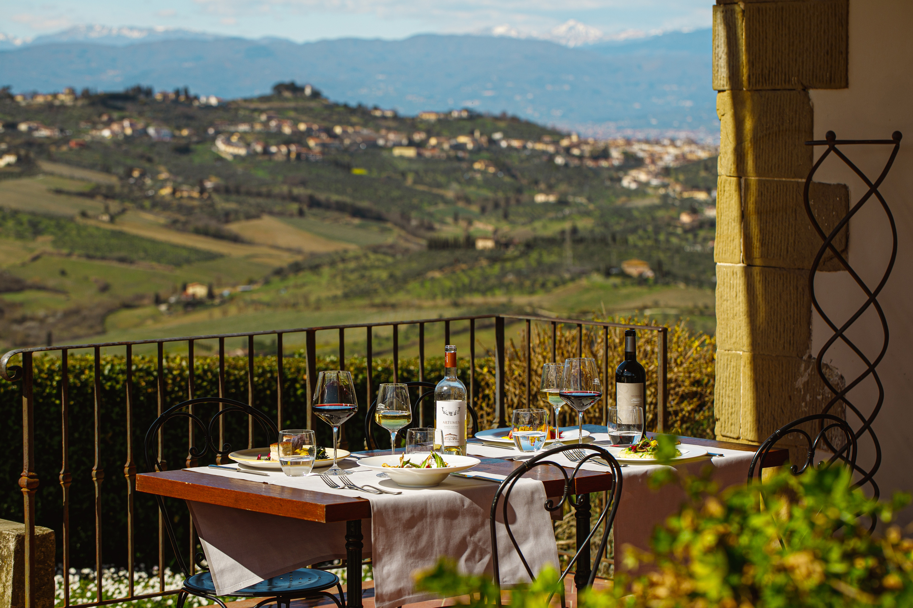 Scenic Tuscan terrace dining with wine, overlooking rolling hills and distant mountains.