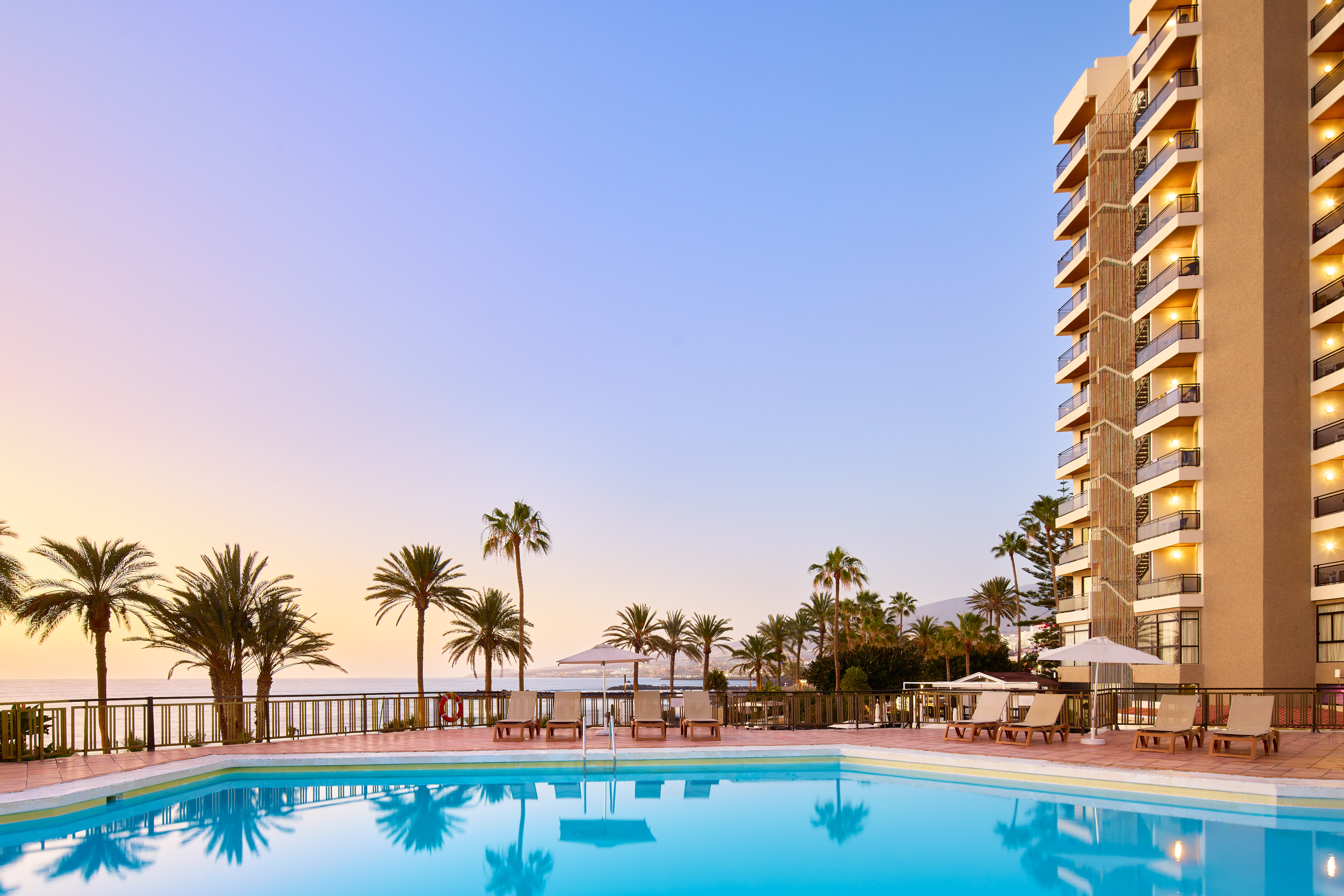 Resort swimming pool with palm trees, ocean view, and hotel at sunset.