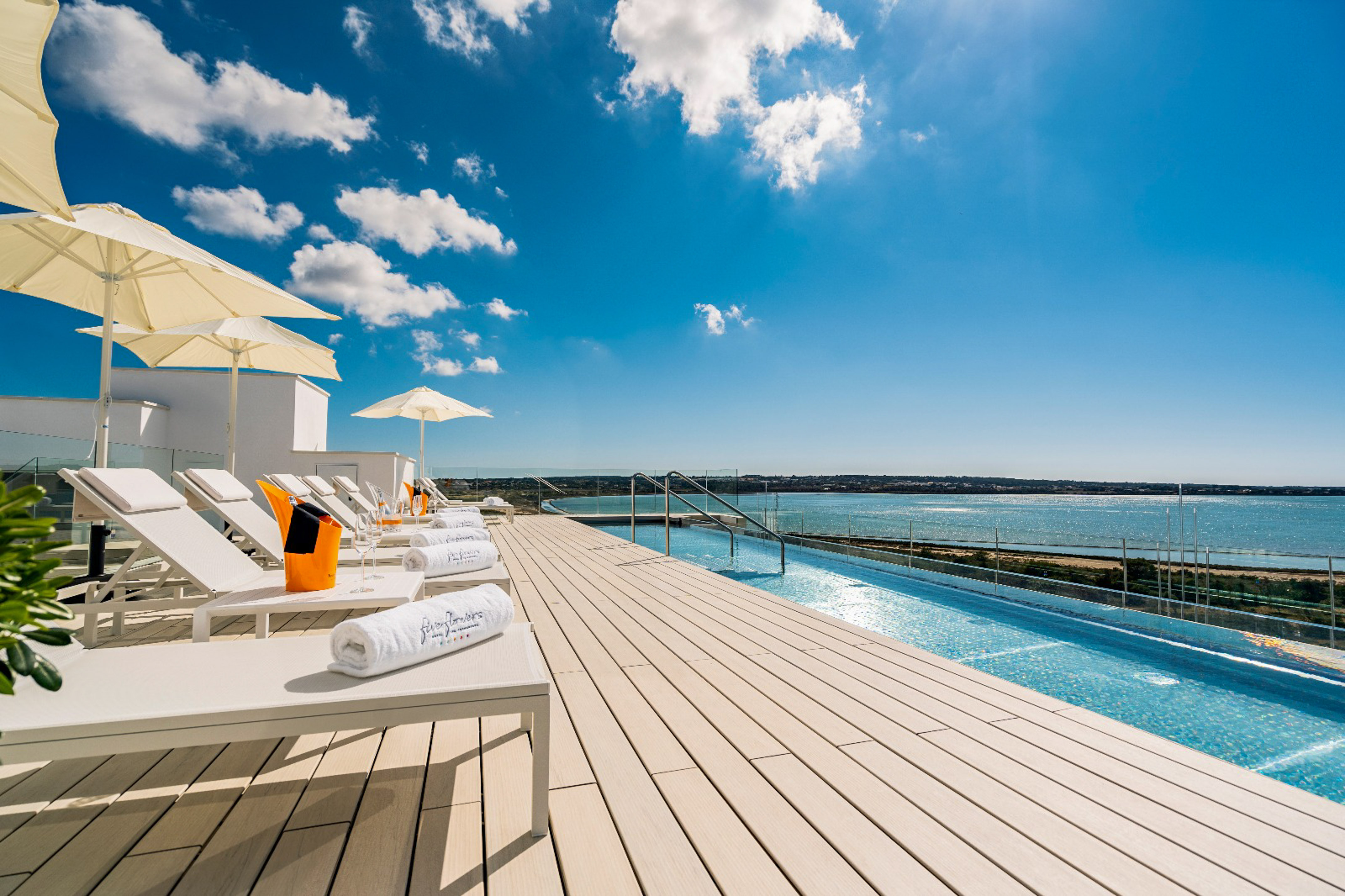Sunny hotel rooftop infinity pool overlooking ocean, with white sun loungers.