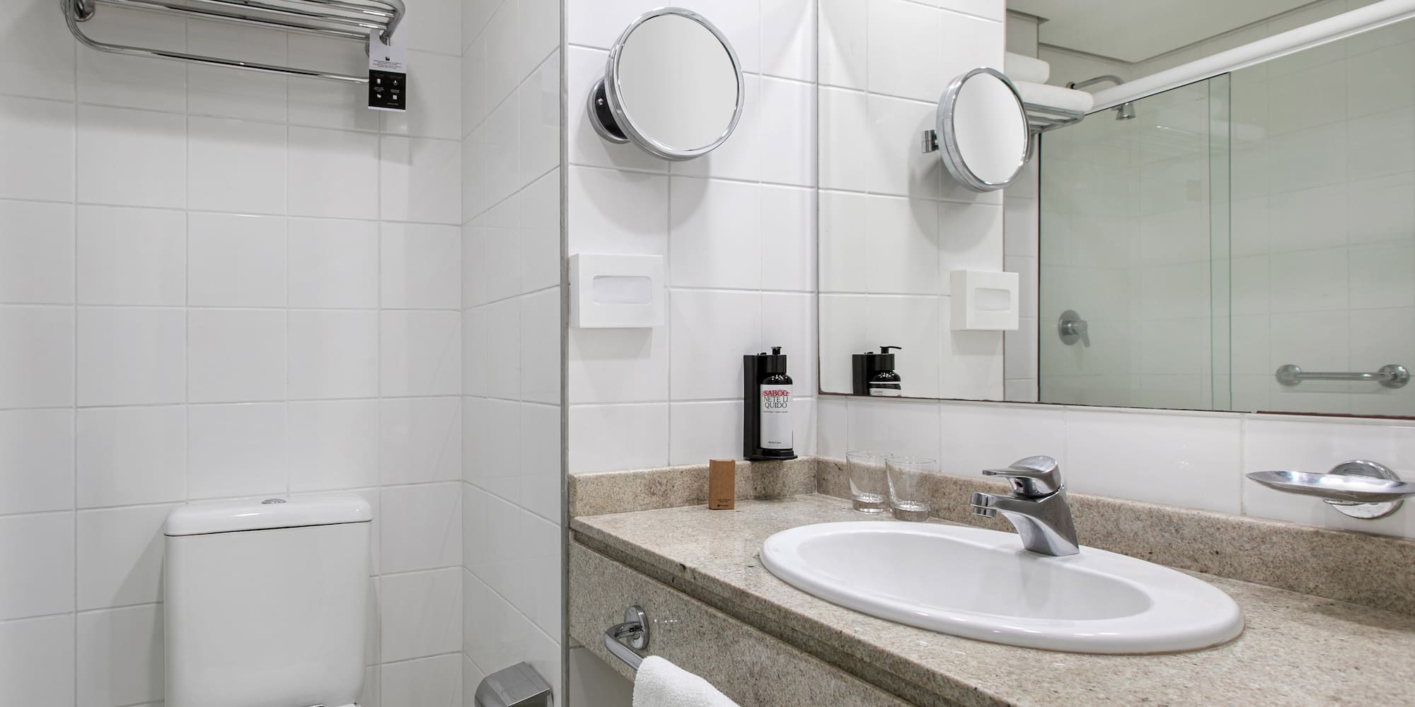 White tiled hotel bathroom with vanity, sink, toilet, and shower.