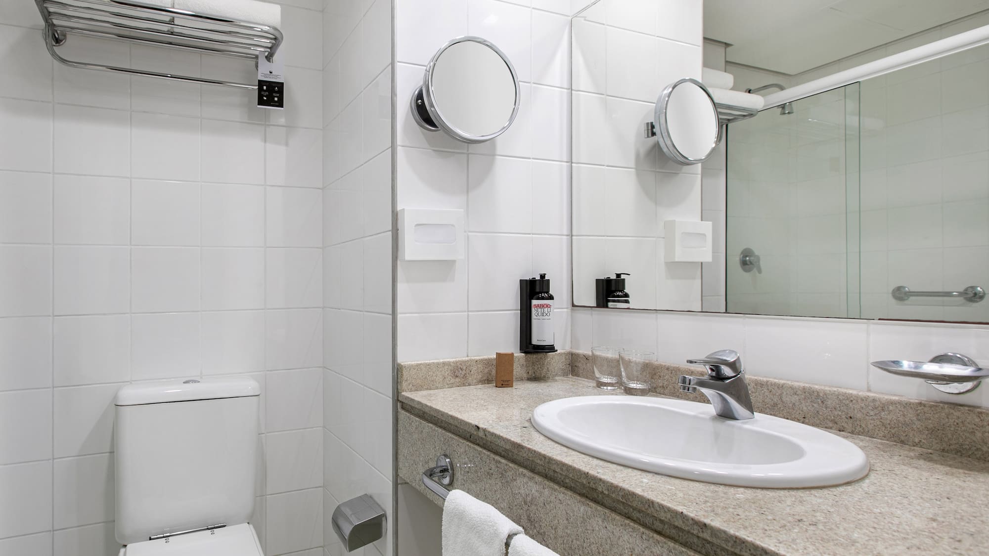 White tiled hotel bathroom with vanity, sink, toilet, and shower.