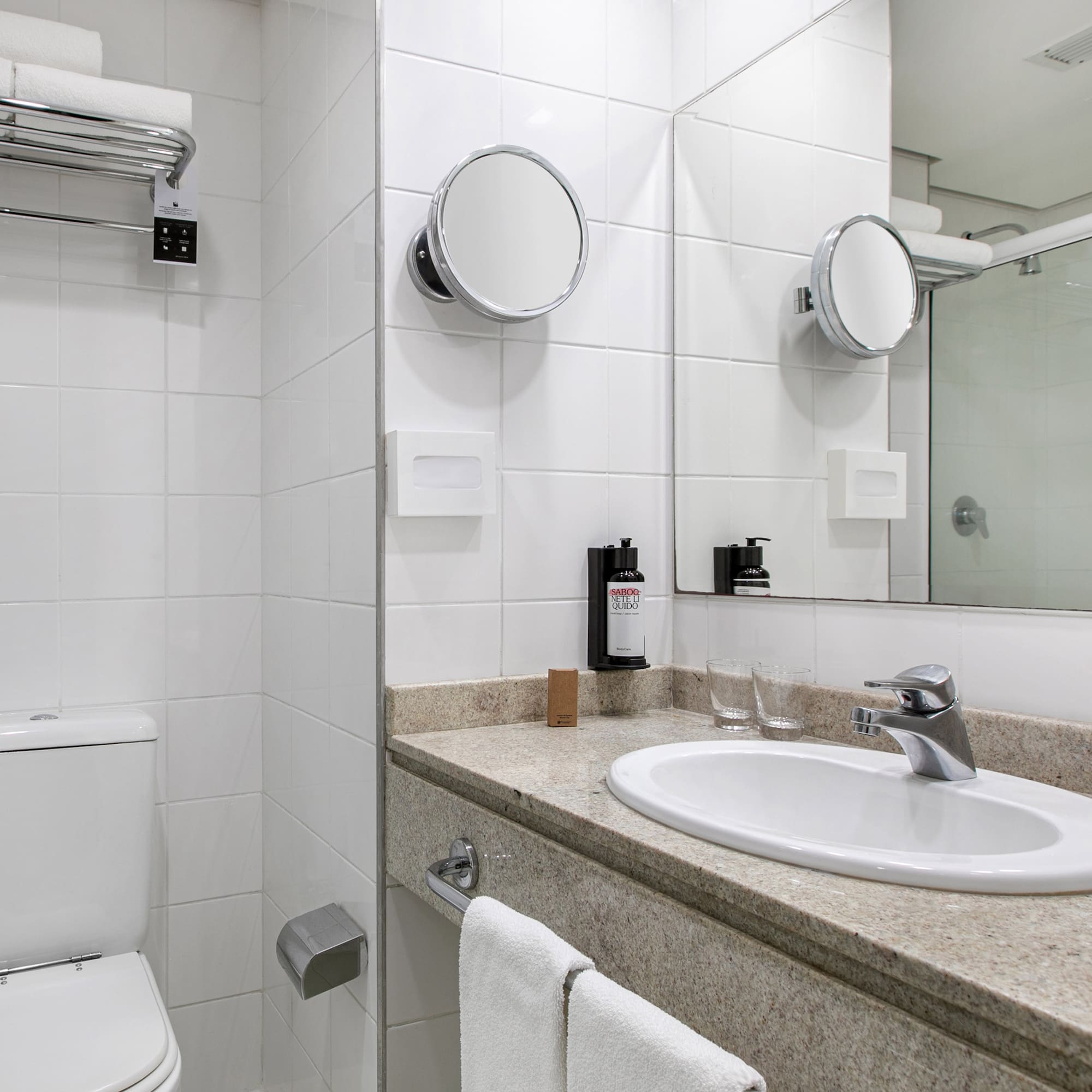 White tiled hotel bathroom with vanity, sink, toilet, and shower.