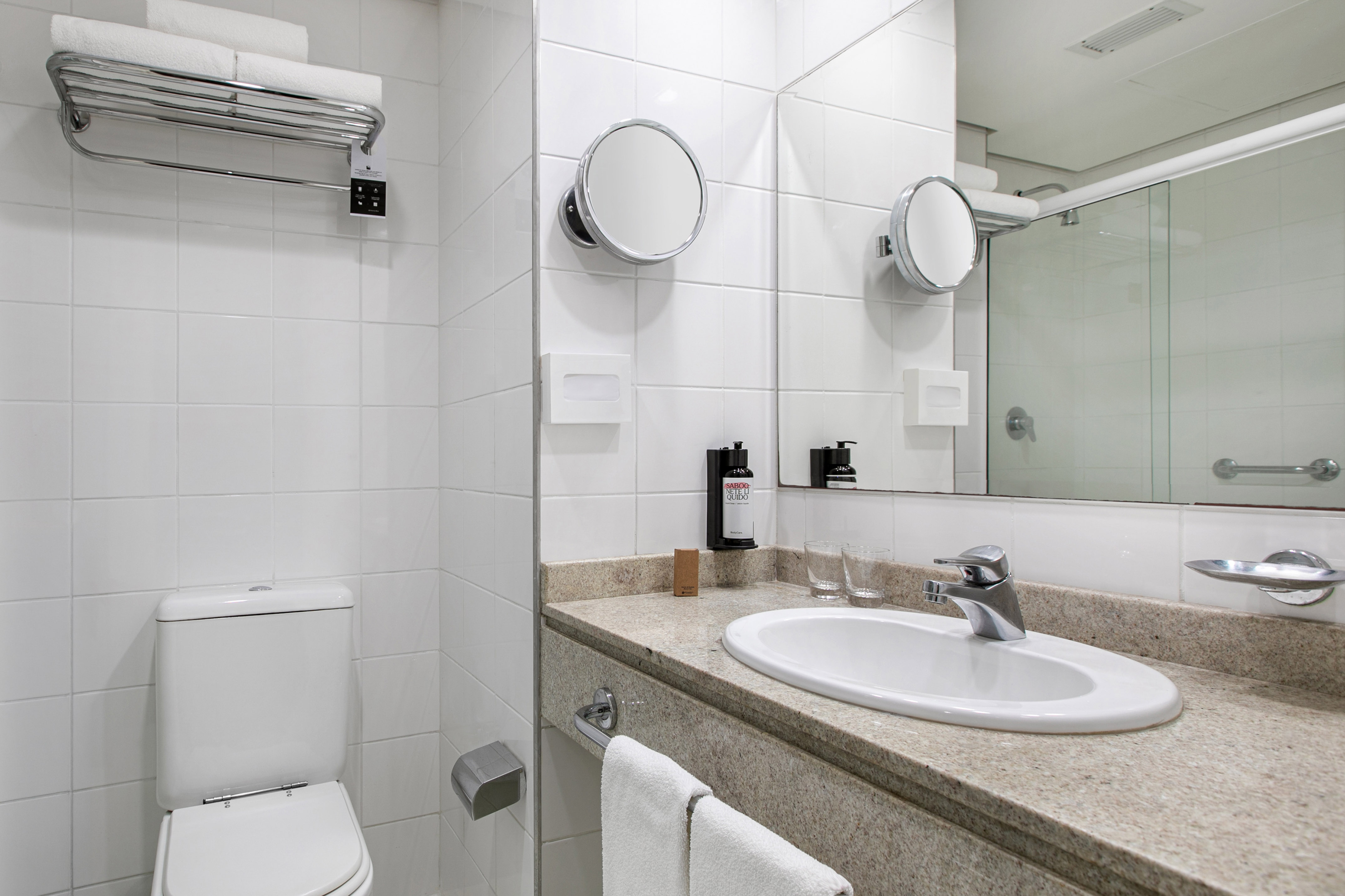 White tiled hotel bathroom with vanity, sink, toilet, and shower.