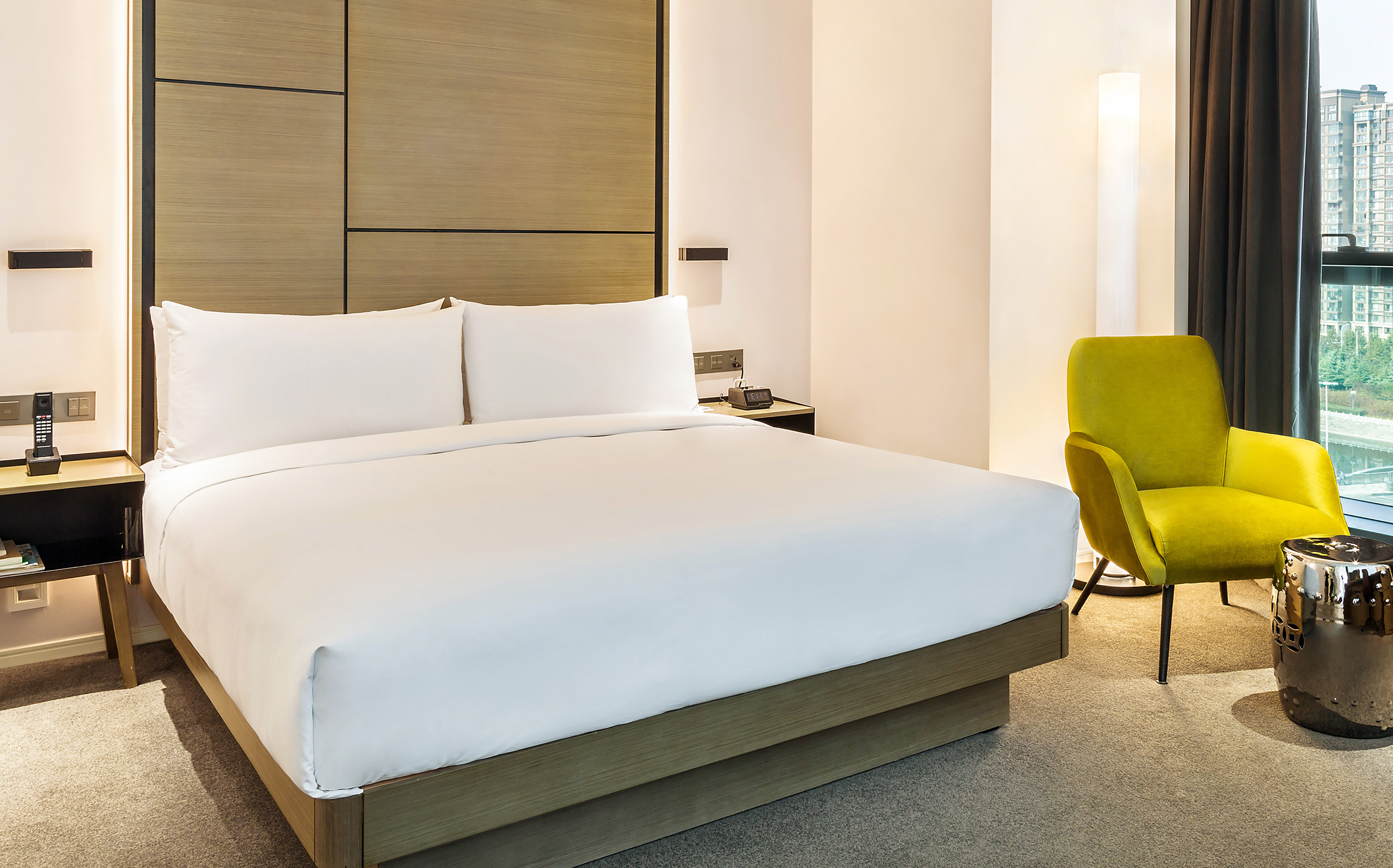 Modern hotel room: king bed, wooden headboard, lime green armchair, city view.