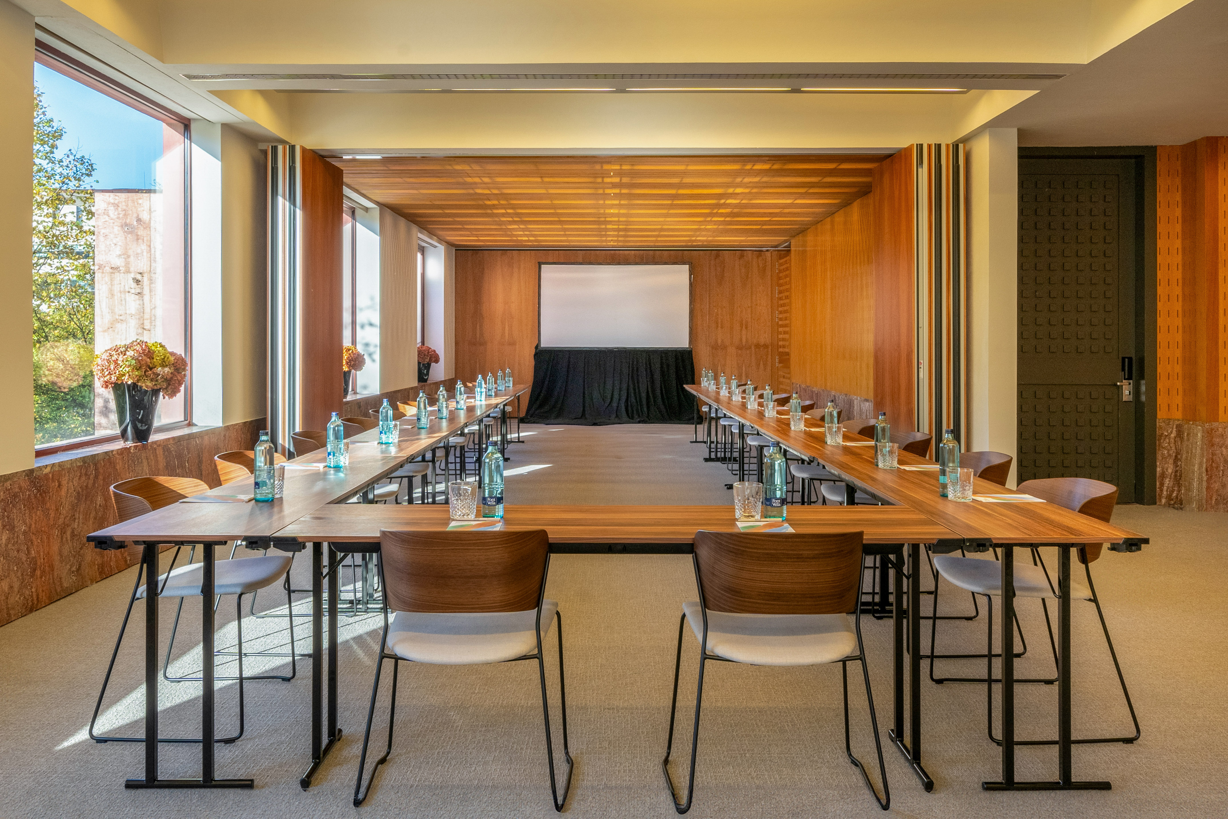 U-shaped conference room with wooden tables, modern chairs, windows, and projector screen.