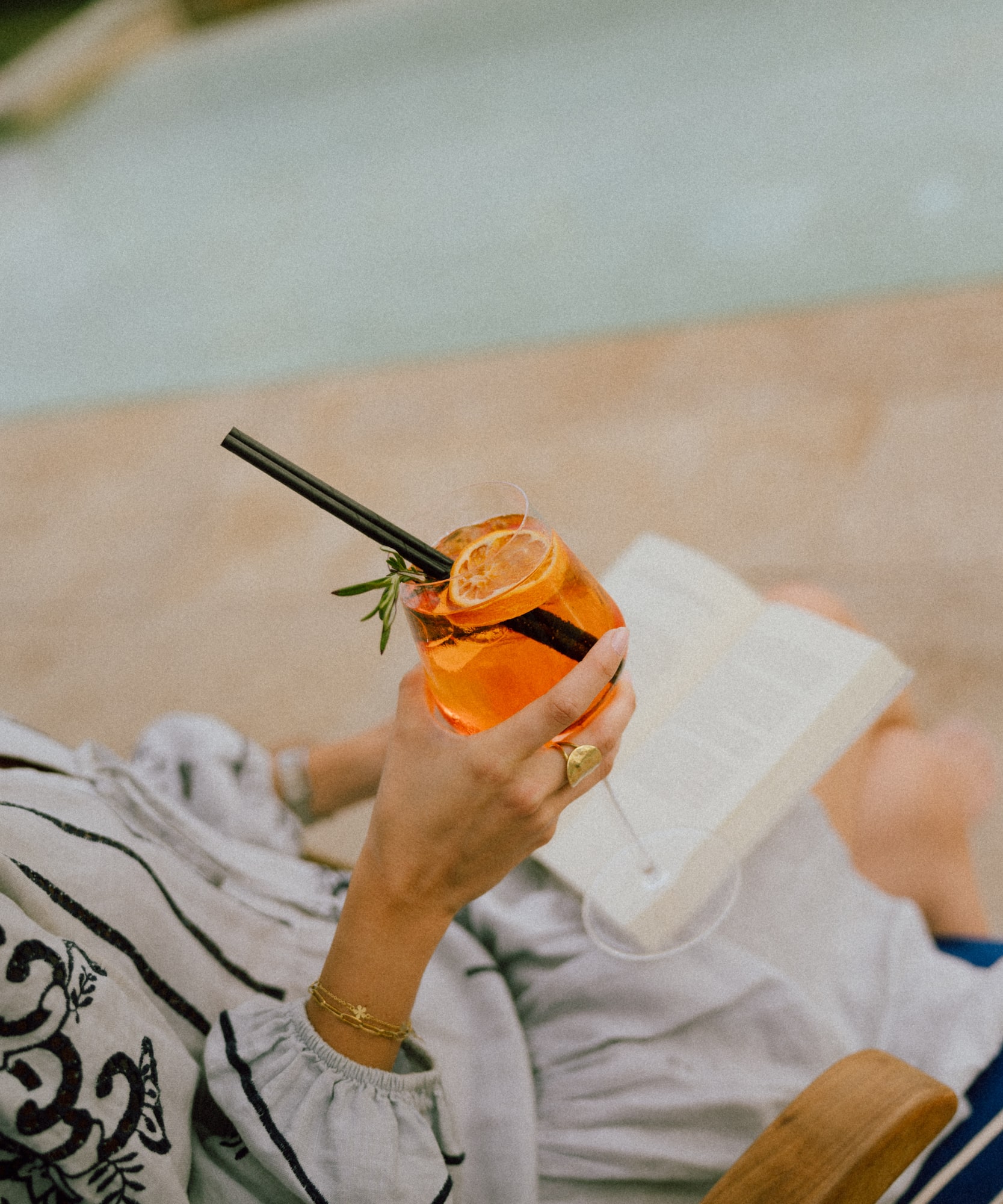 Woman's hand holding Aperol Spritz cocktail, reading by the pool.