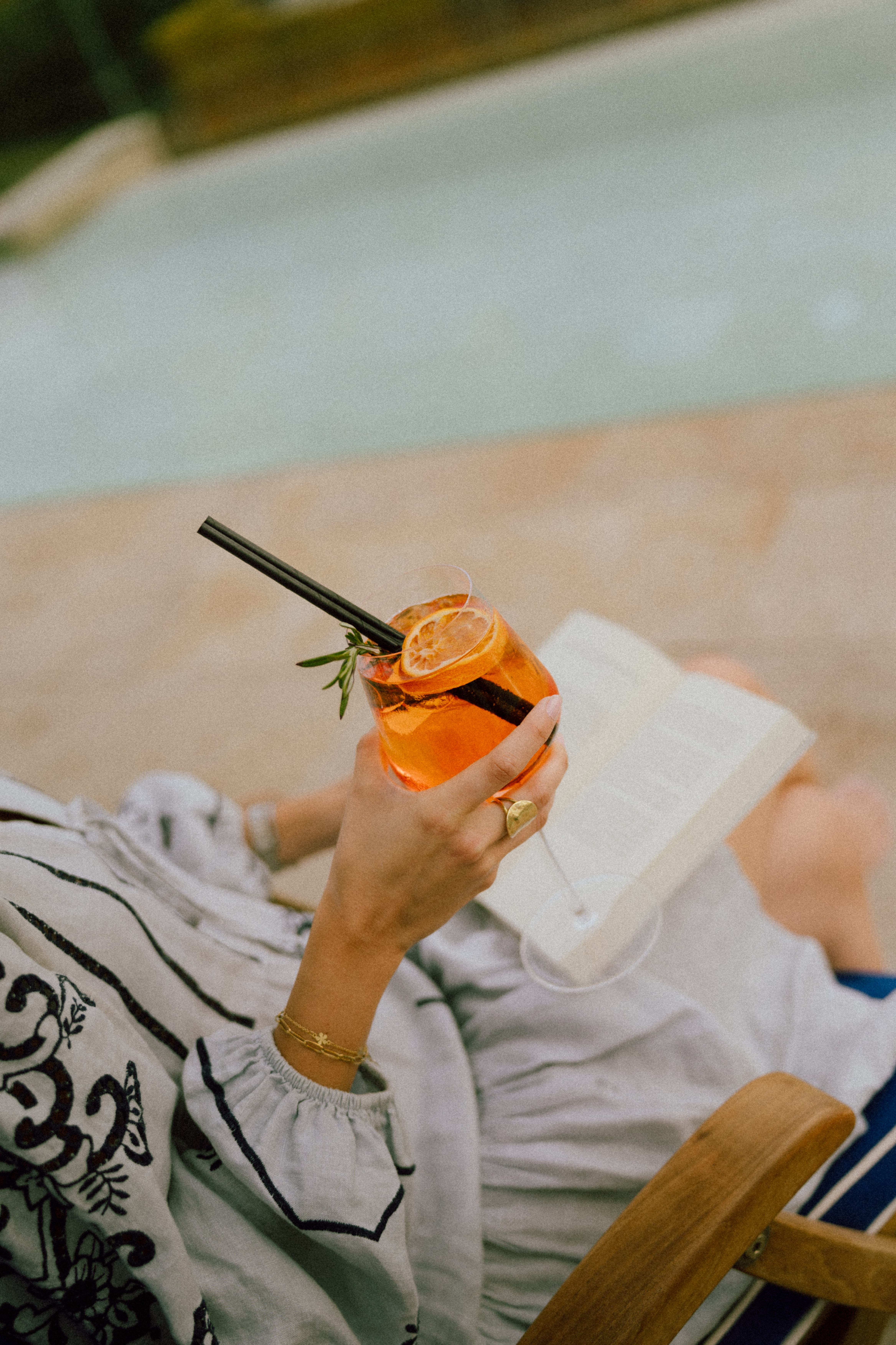 Woman's hand holding Aperol Spritz cocktail, reading by the pool.