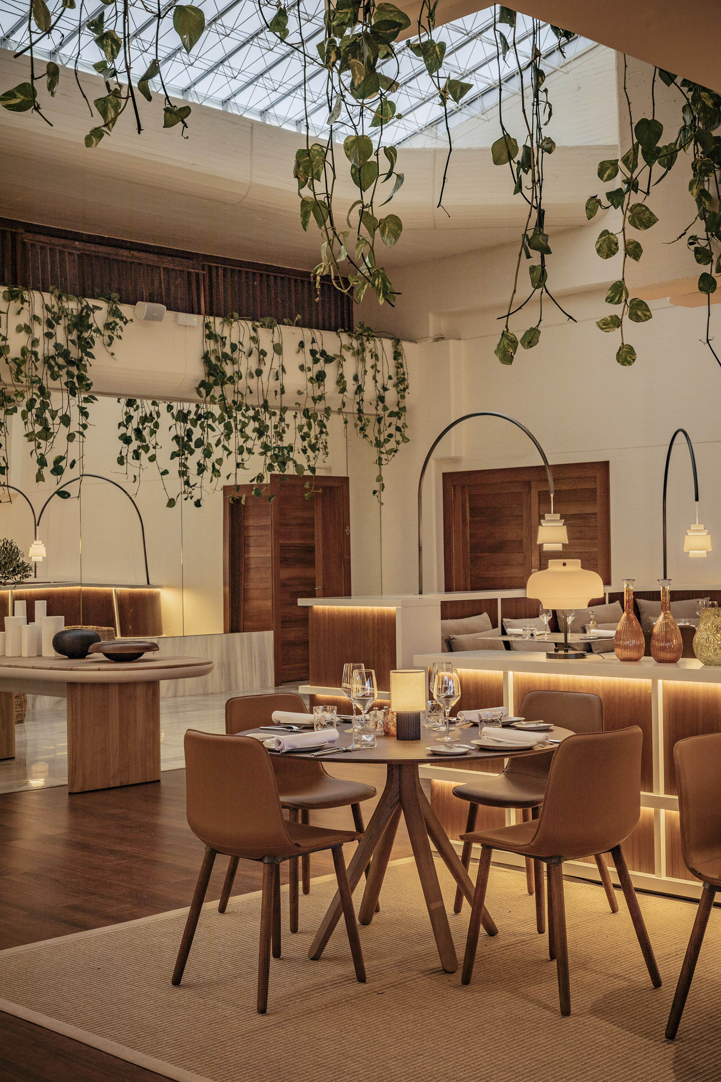 Modern restaurant dining with hanging plants, glass roof, and warm lighting.