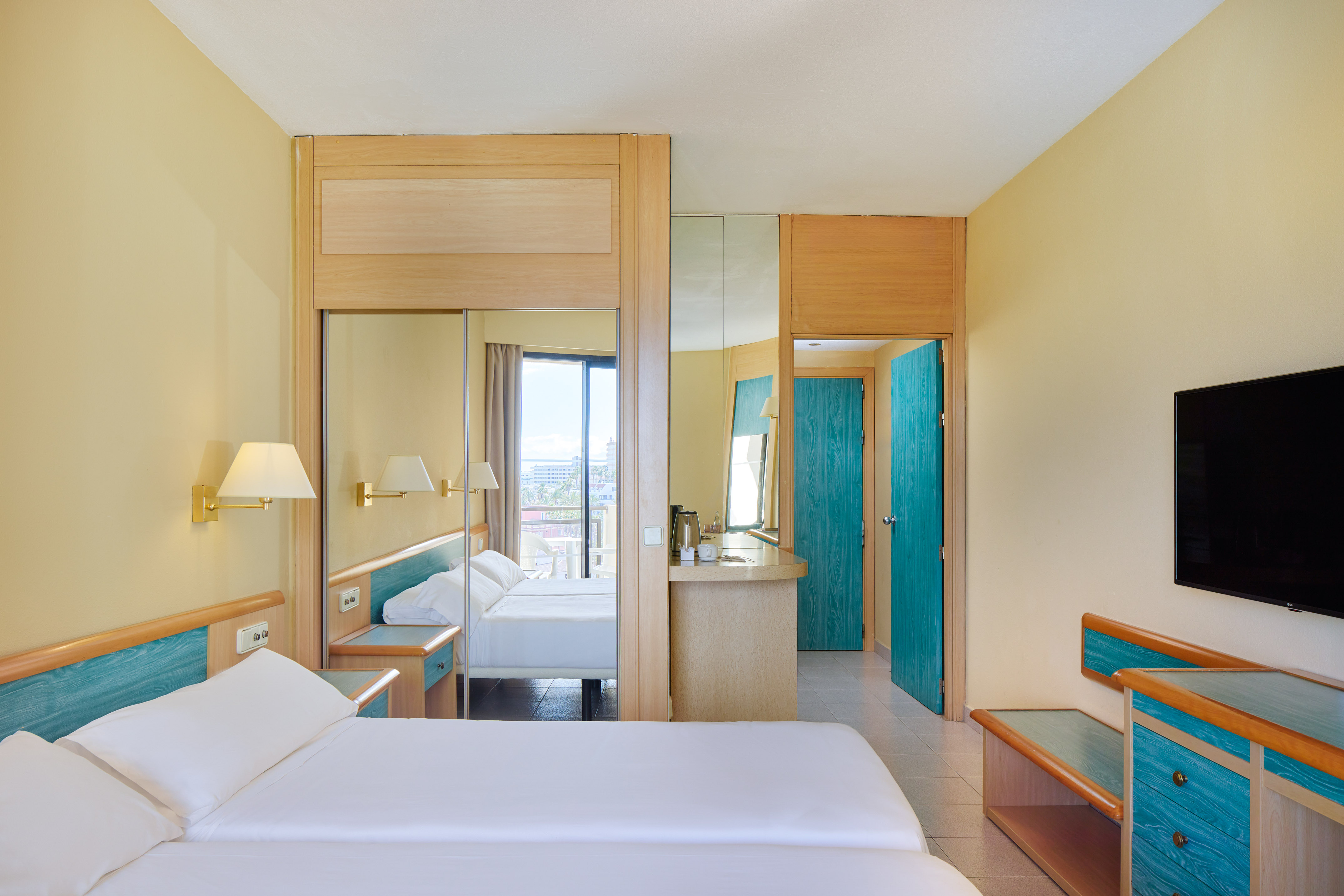 Modern hotel room with twin beds, yellow walls, teal accents, and balcony.