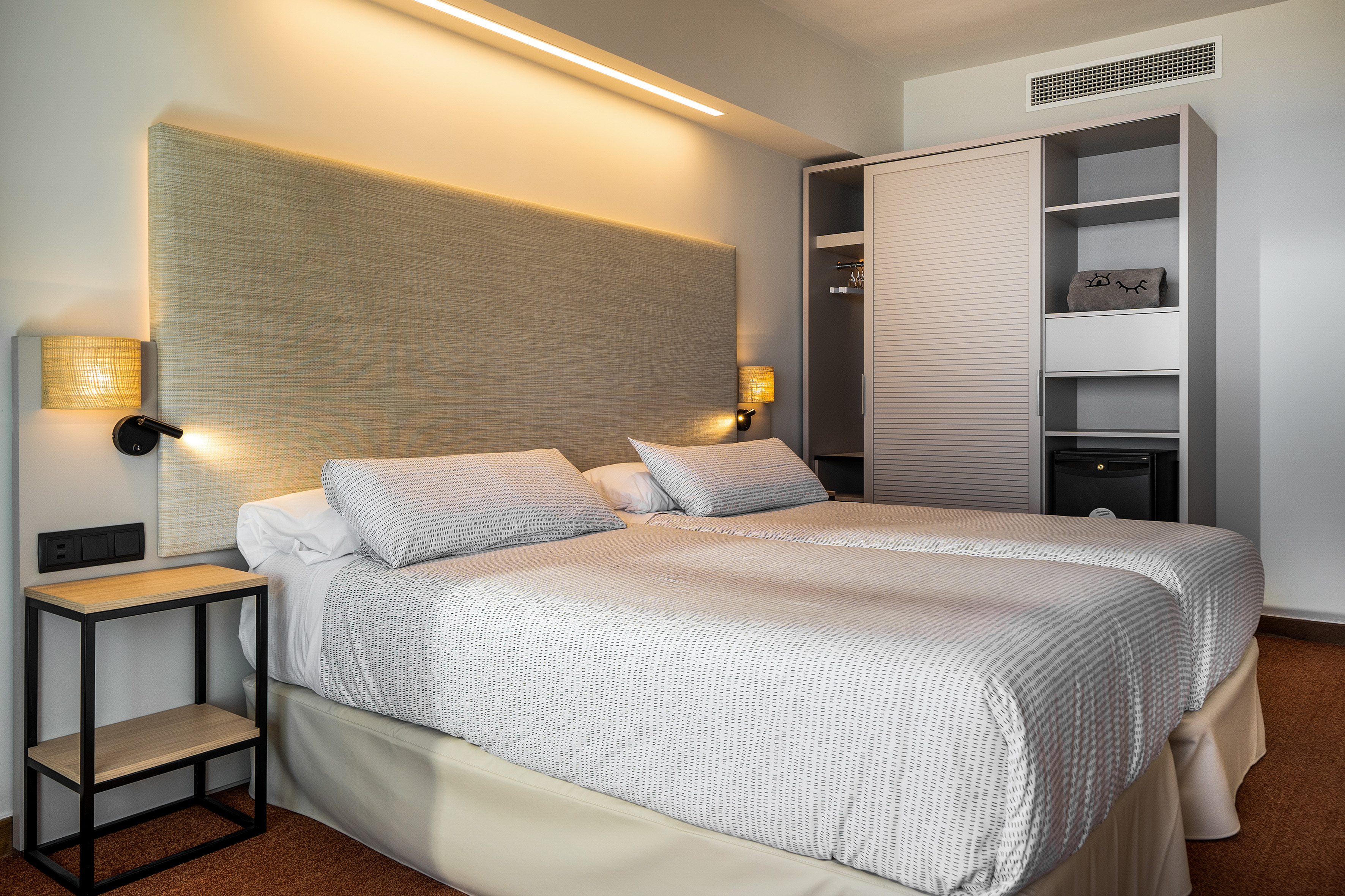 Modern hotel room with twin beds, backlit headboard, and wardrobe.