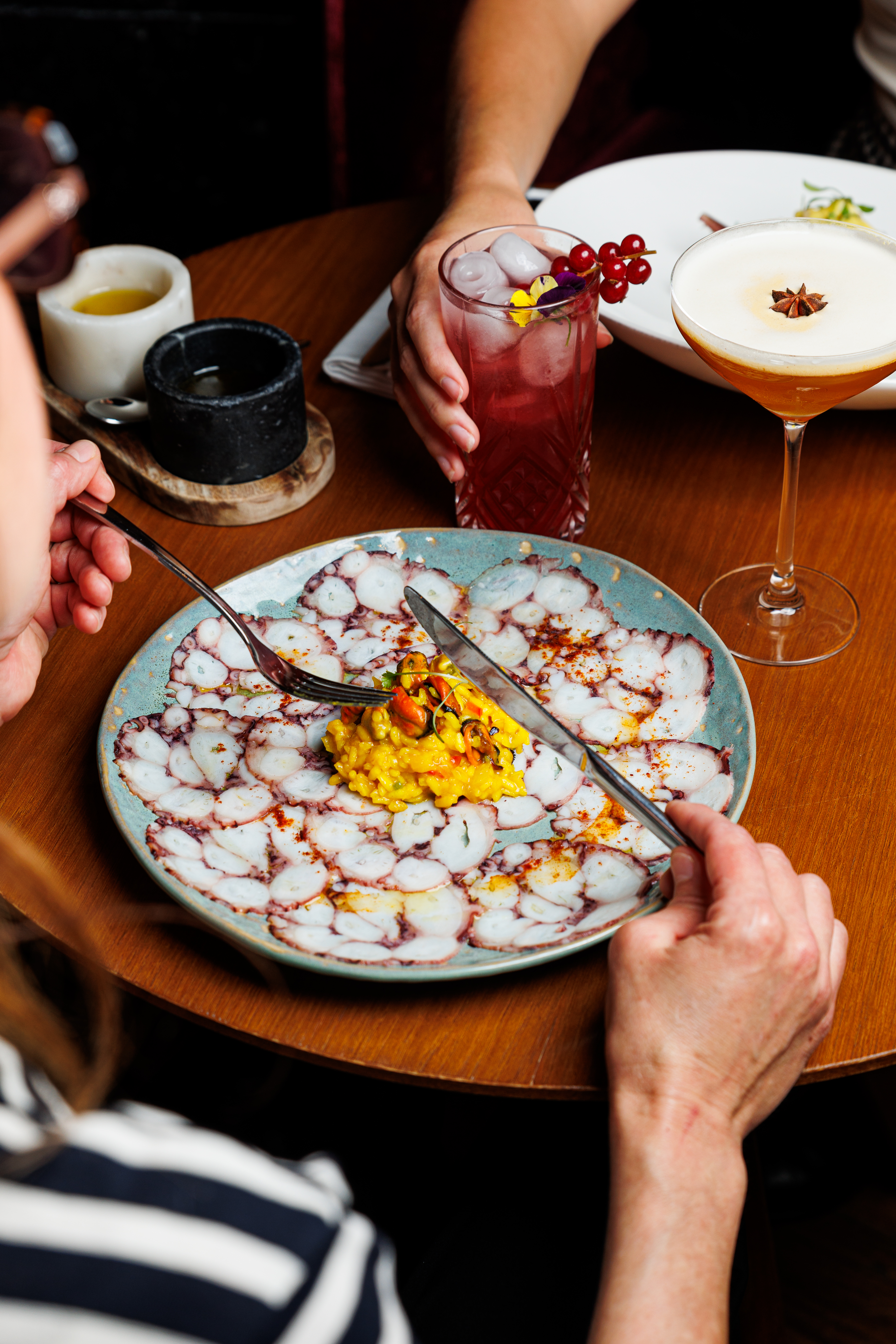 Octopus carpaccio and seafood risotto on a plate, with cocktails at a restaurant.