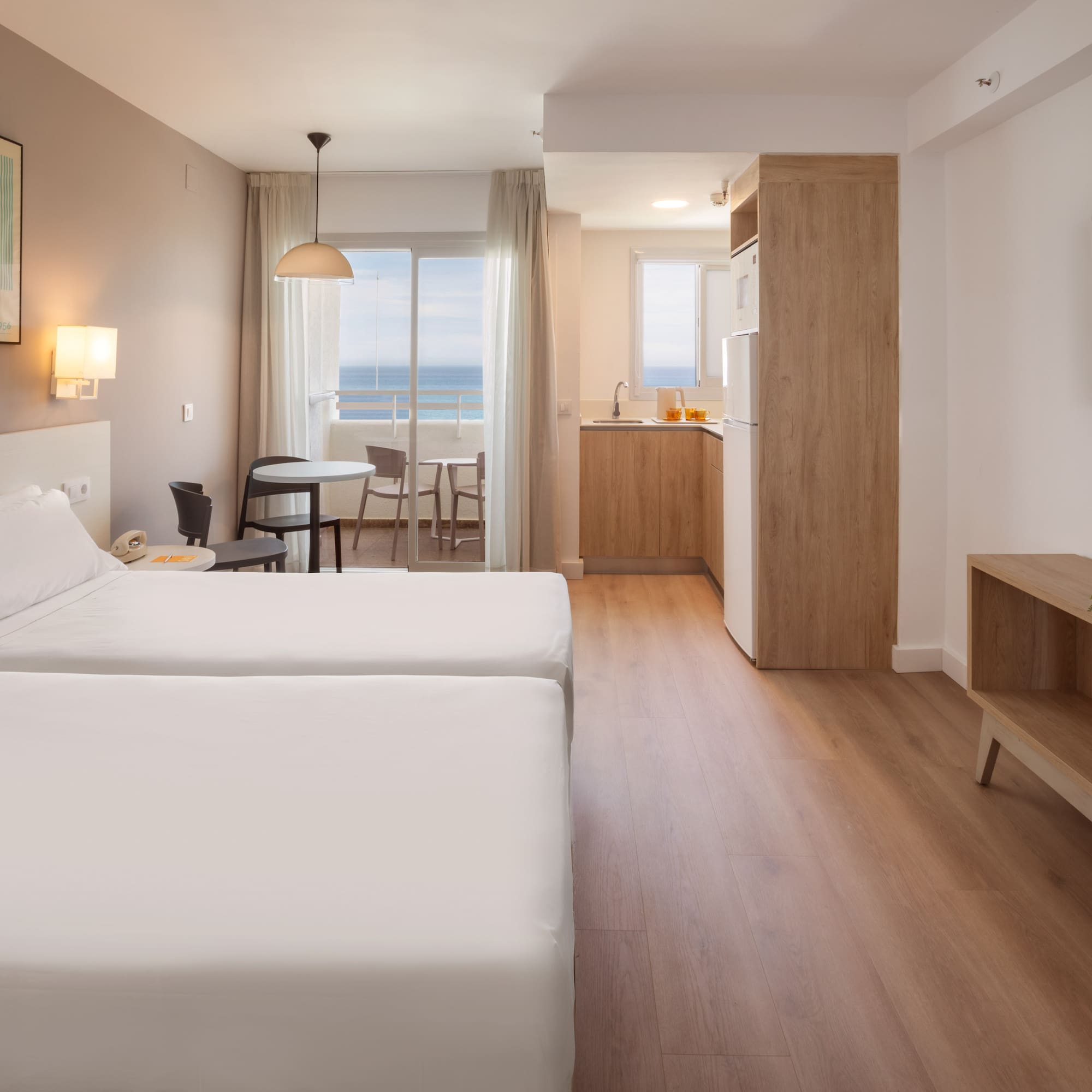 Modern hotel studio apartment with twin beds, kitchenette, balcony, and sea view.