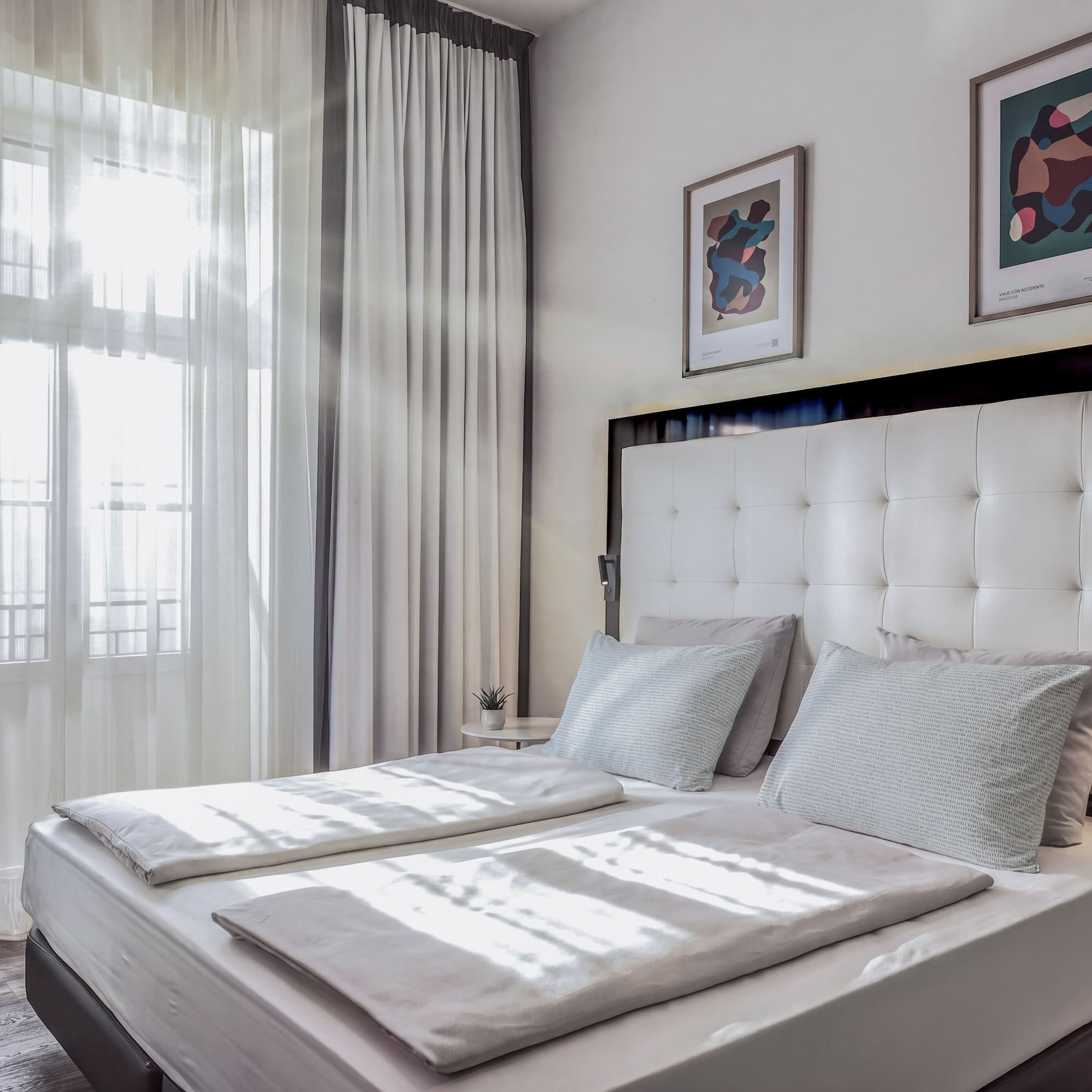 Bright hotel bedroom with white double bed, sheer curtains, and modern art.