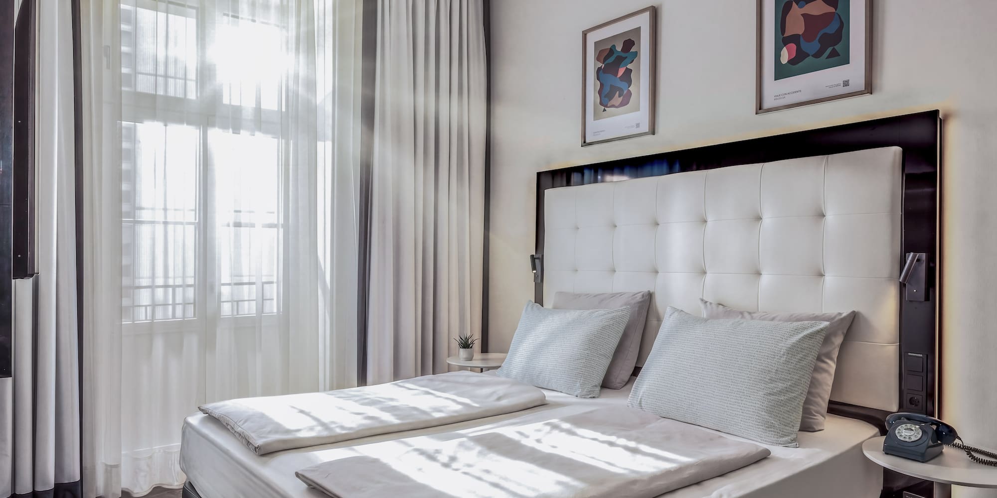 Bright hotel bedroom with white double bed, sheer curtains, and modern art.