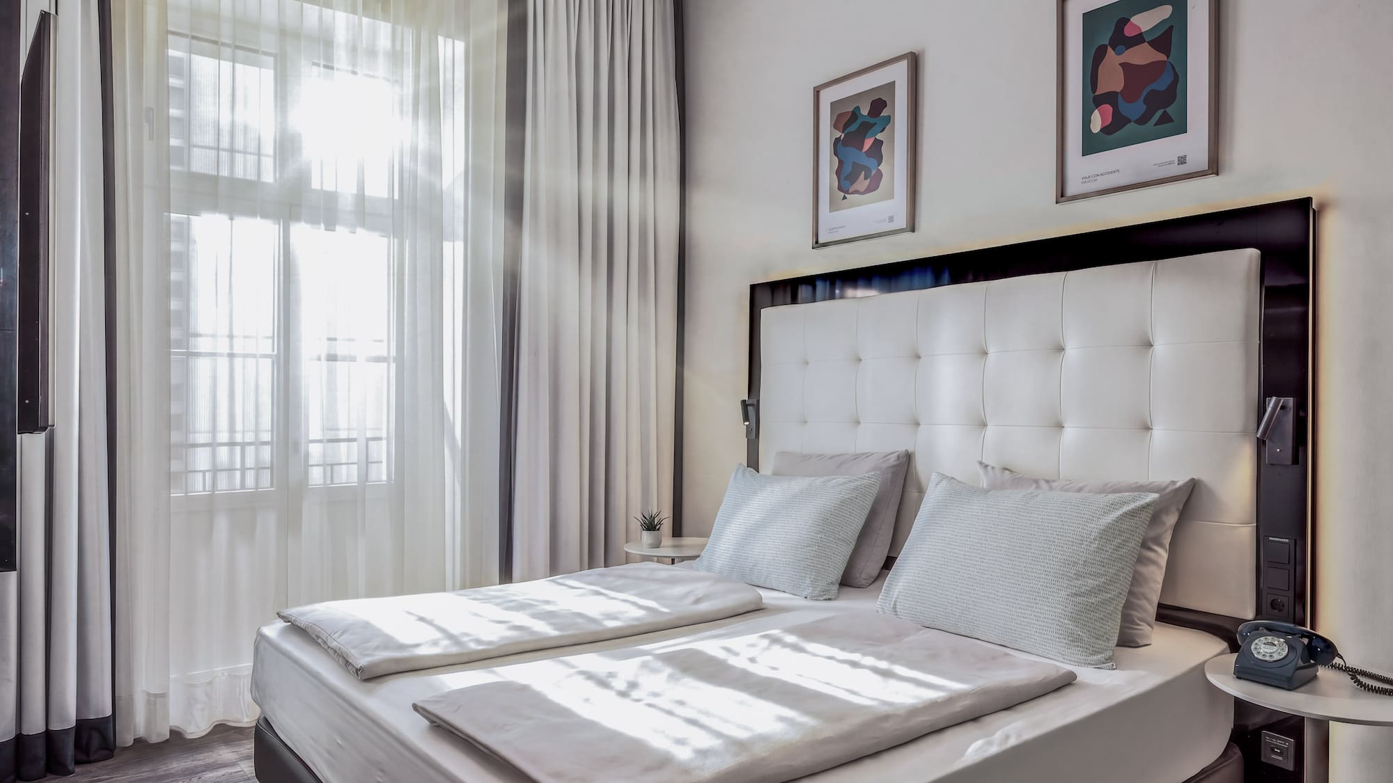 Bright hotel bedroom with white double bed, sheer curtains, and modern art.