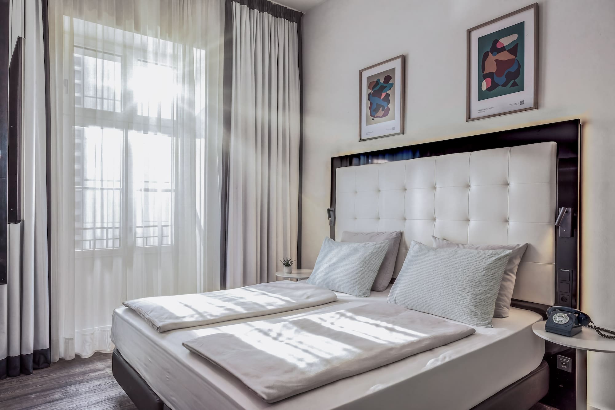 Bright hotel bedroom with white double bed, sheer curtains, and modern art.