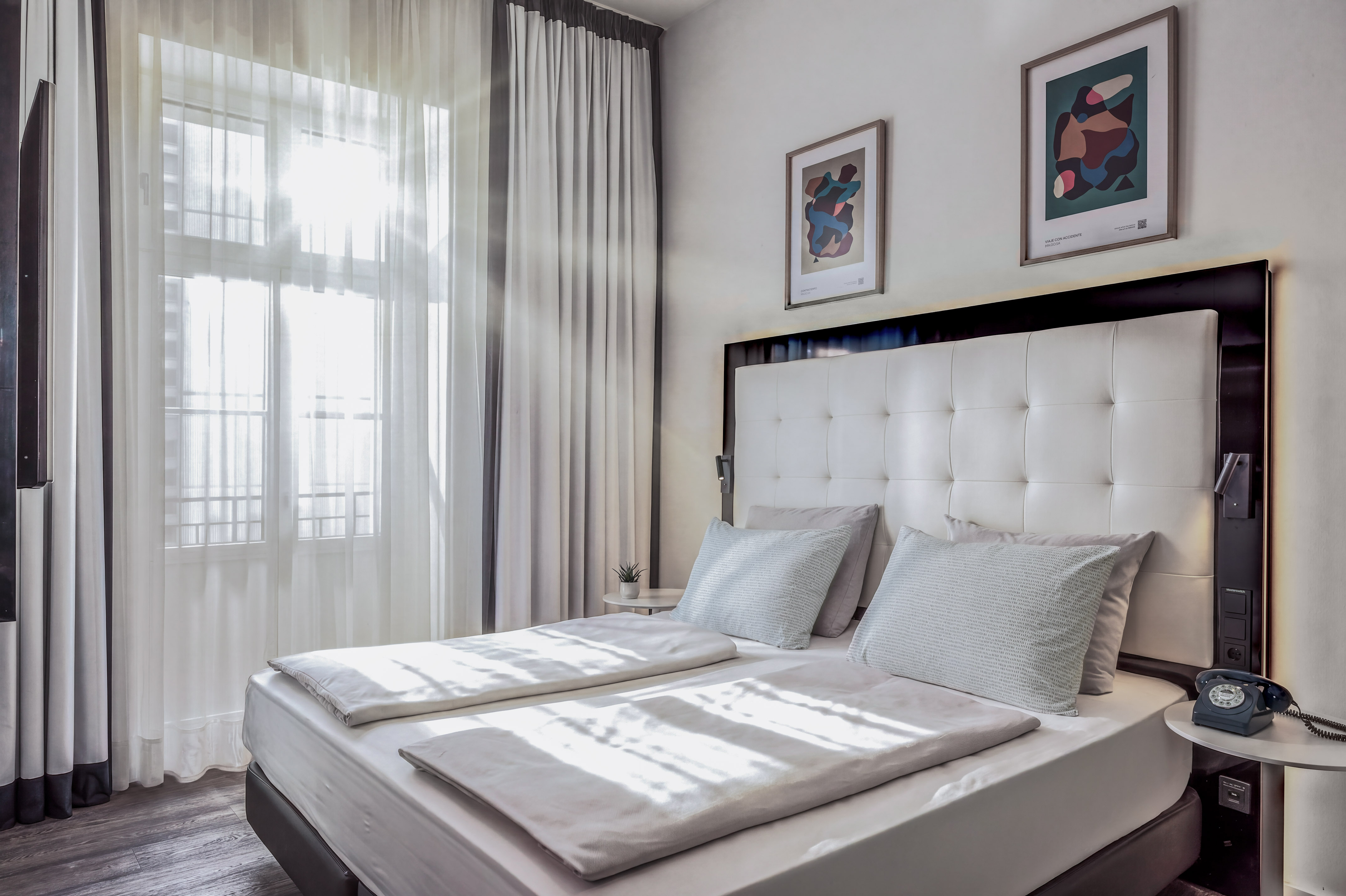 Bright hotel bedroom with white double bed, sheer curtains, and modern art.