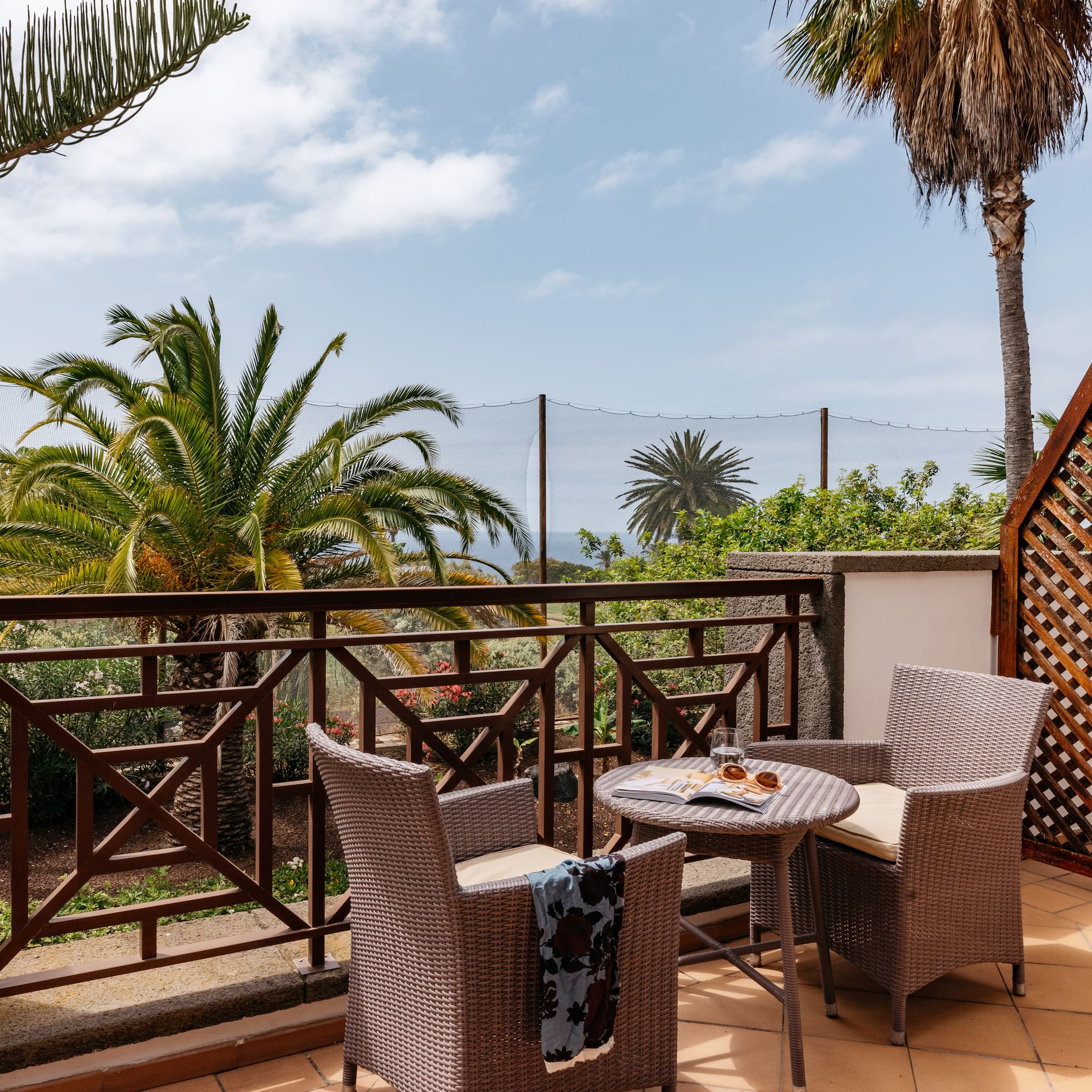 Tropical balcony with wicker furniture, palm trees, and distant ocean view.