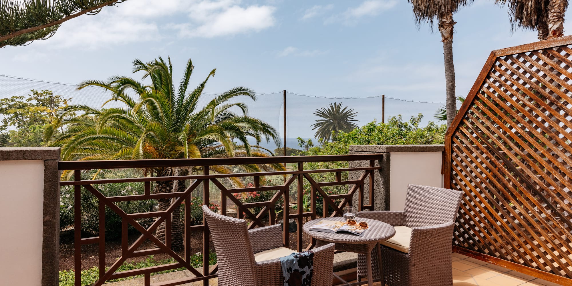 Tropical balcony with wicker furniture, palm trees, and distant ocean view.