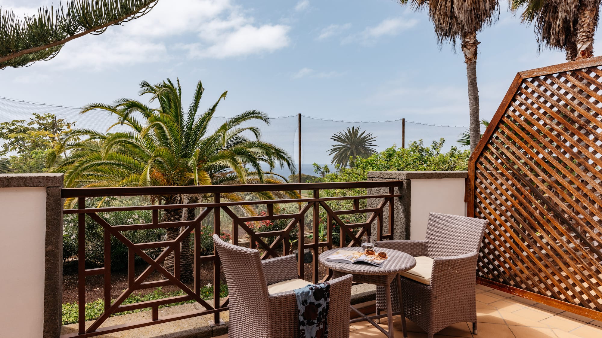 Tropical balcony with wicker furniture, palm trees, and distant ocean view.