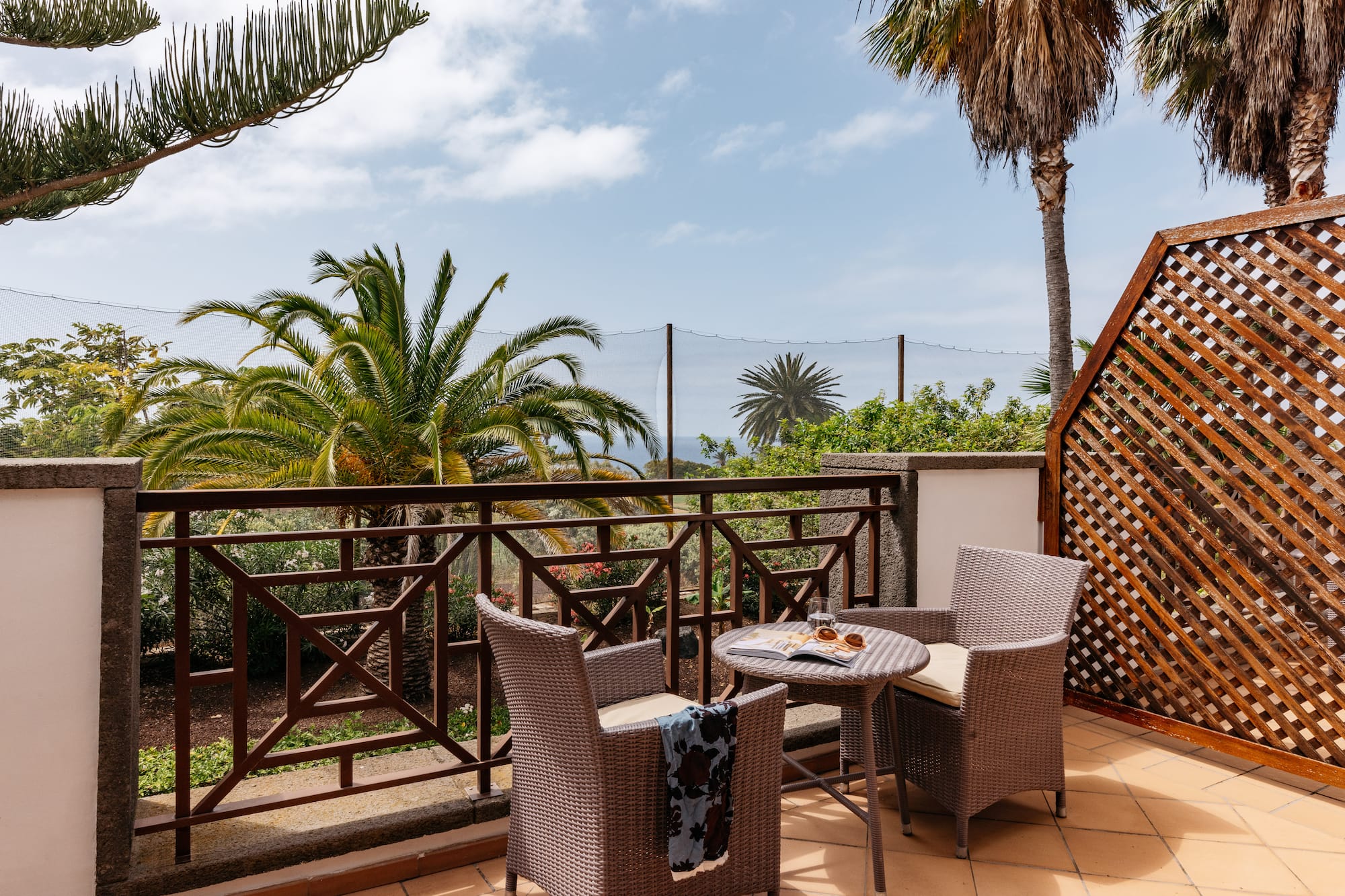Tropical balcony with wicker furniture, palm trees, and distant ocean view.