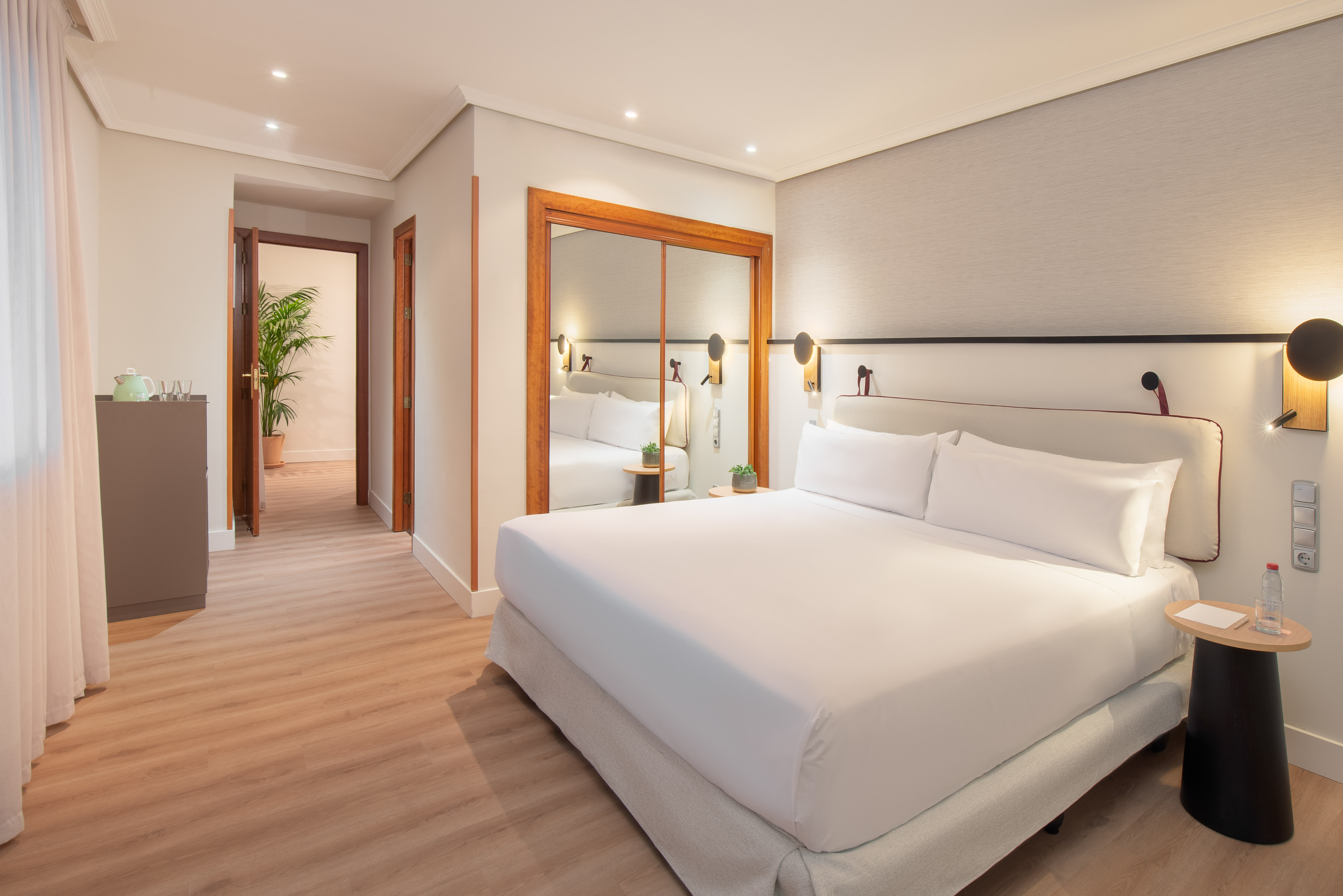Modern hotel bedroom with large white bed, light wood floor, and mirrored closet.