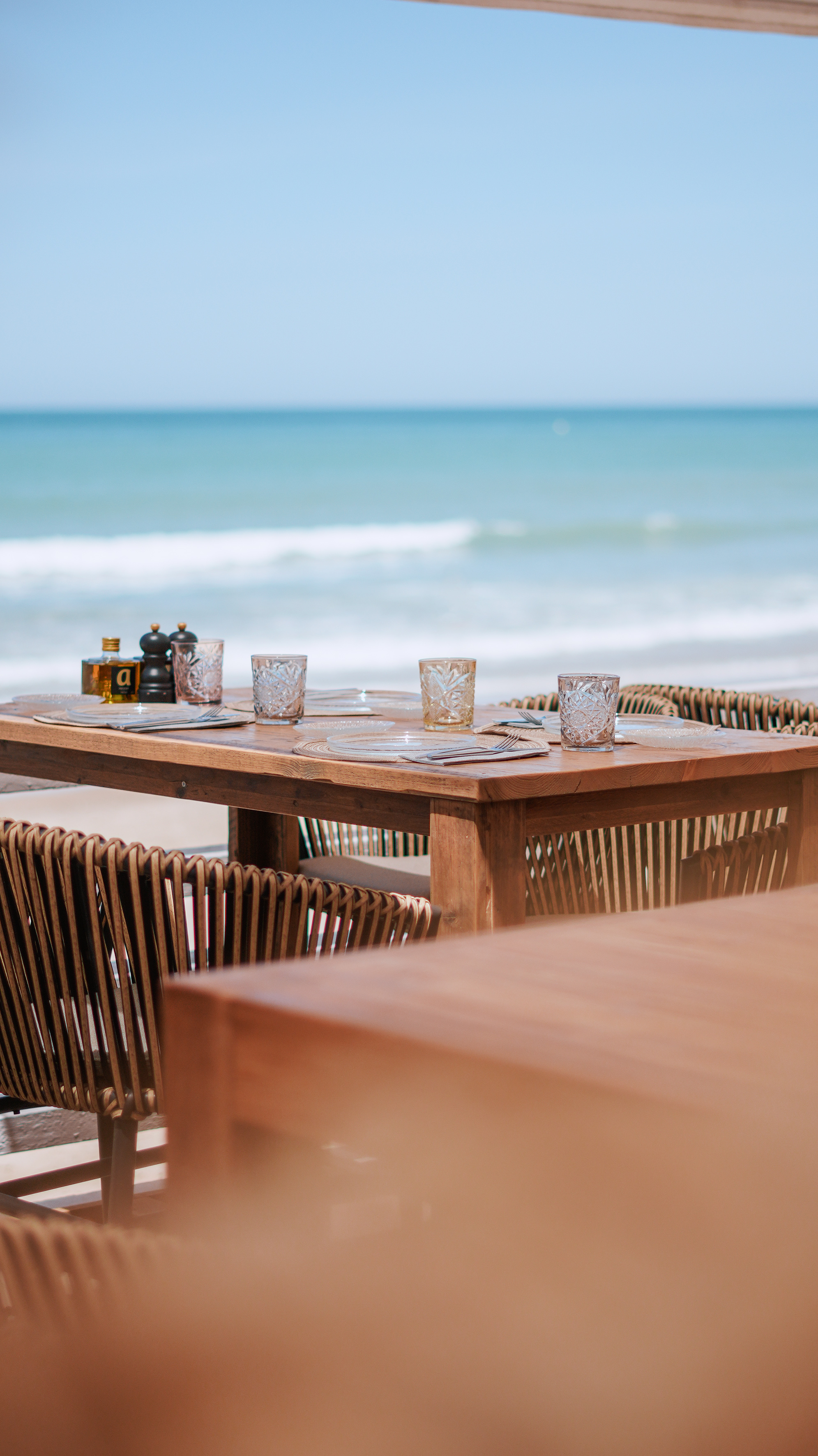 Set wooden dining table and woven chairs at beachfront restaurant with ocean view.