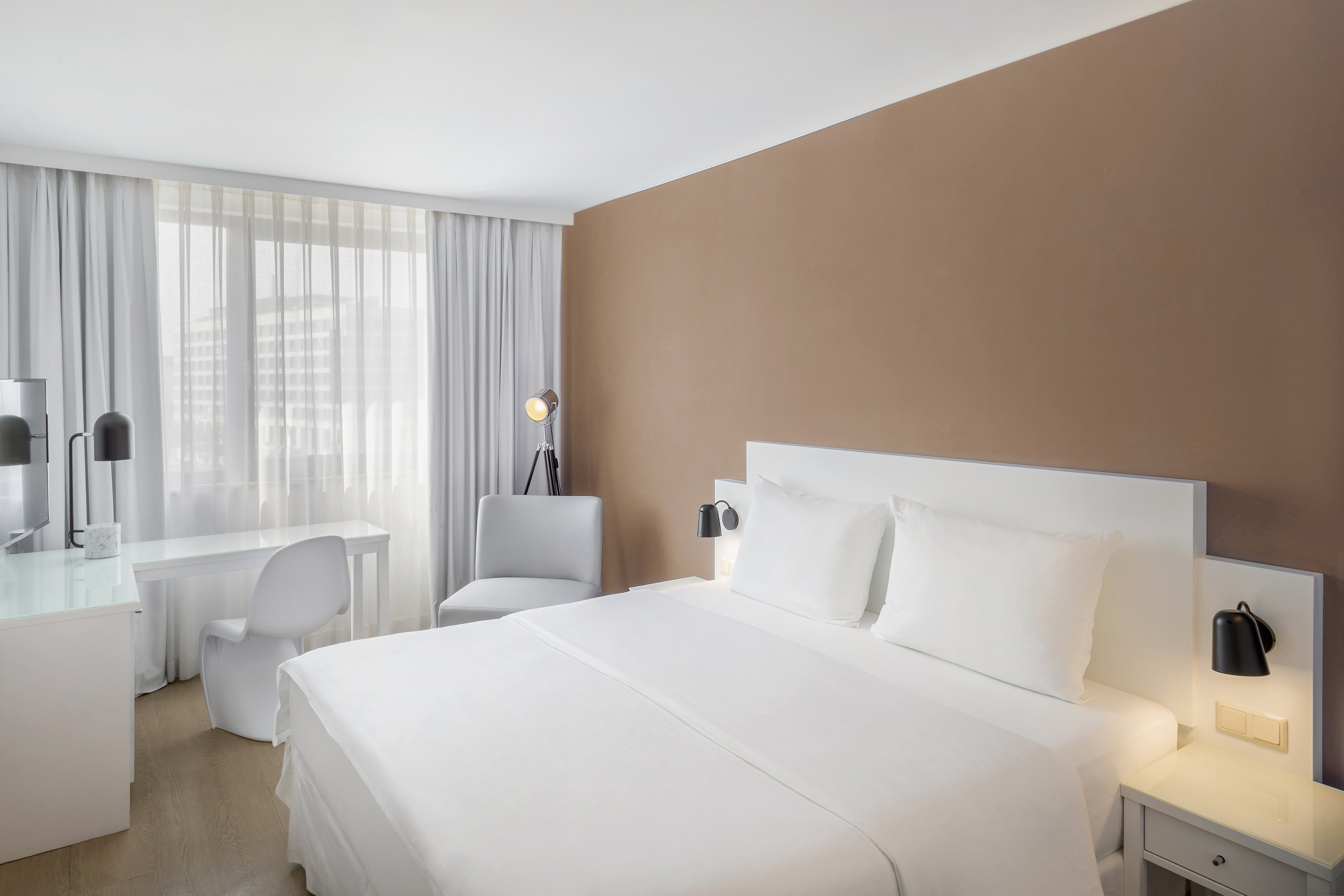 Modern hotel guest room with white bed, brown wall, desk, and large window.