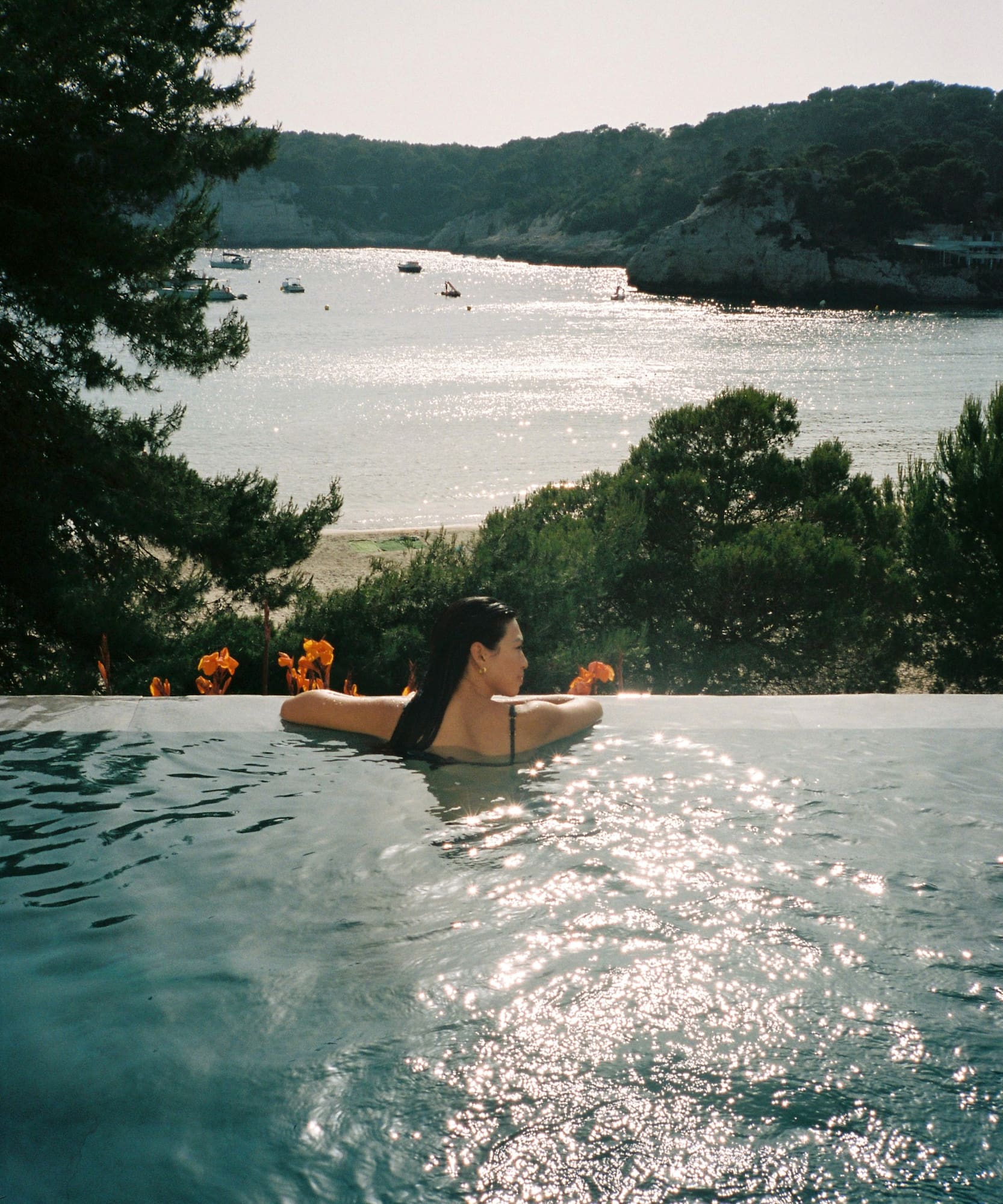 Mujer relajándose en piscina infinita con vista a bahía soleada y montañas.