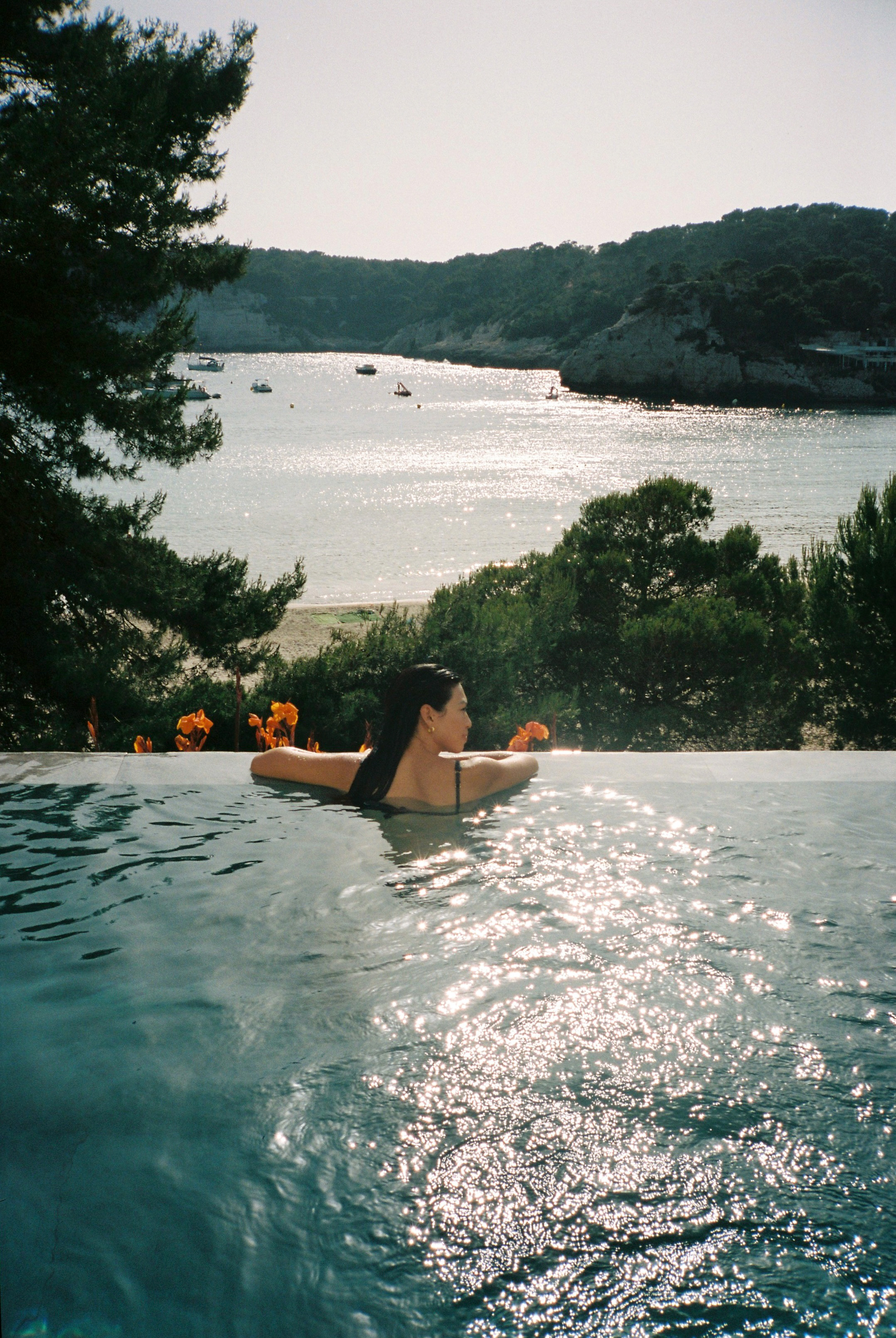 Mujer relajándose en piscina infinita con vista a bahía soleada y montañas.