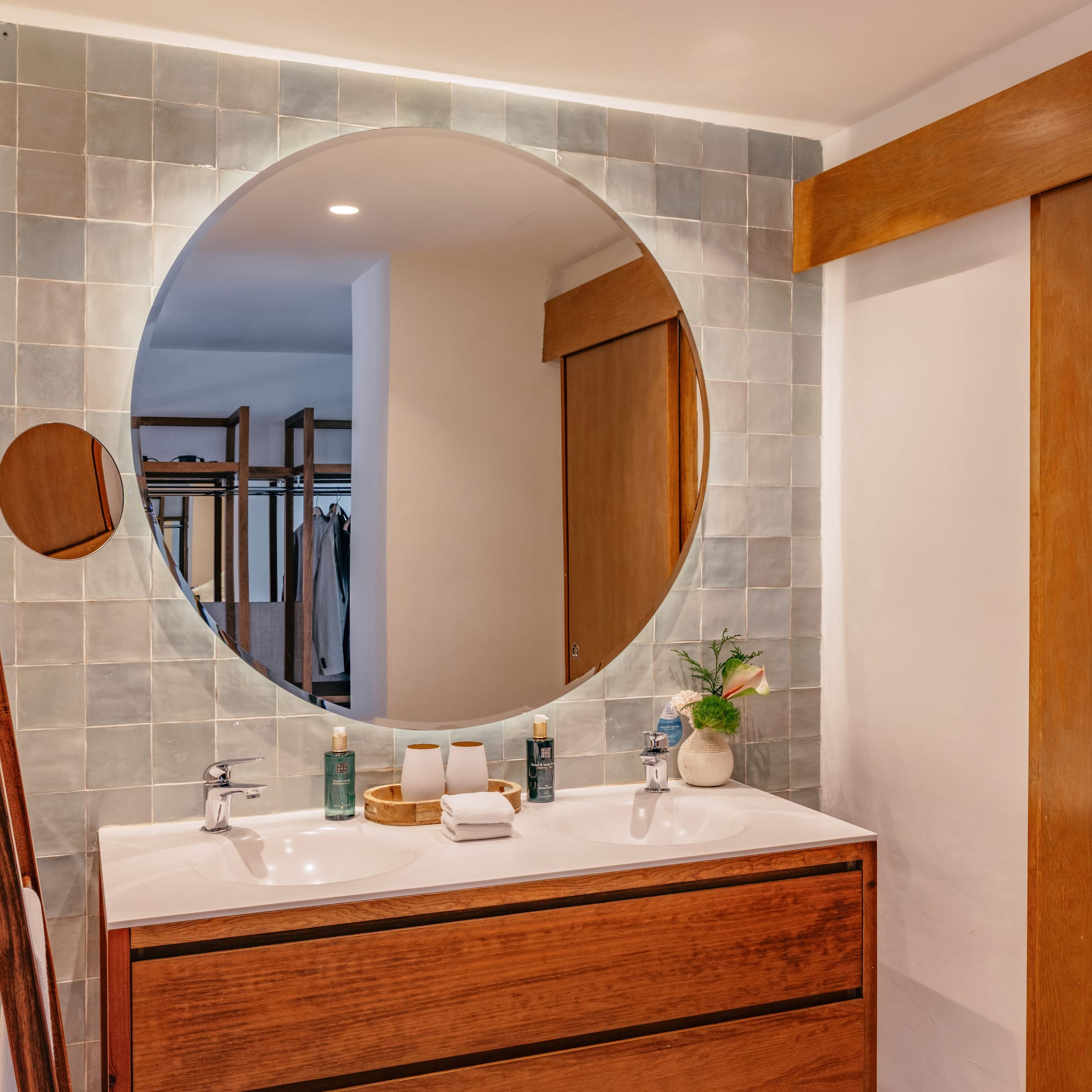 Double sink bathroom vanity with round backlit mirror and grey tiled wall.