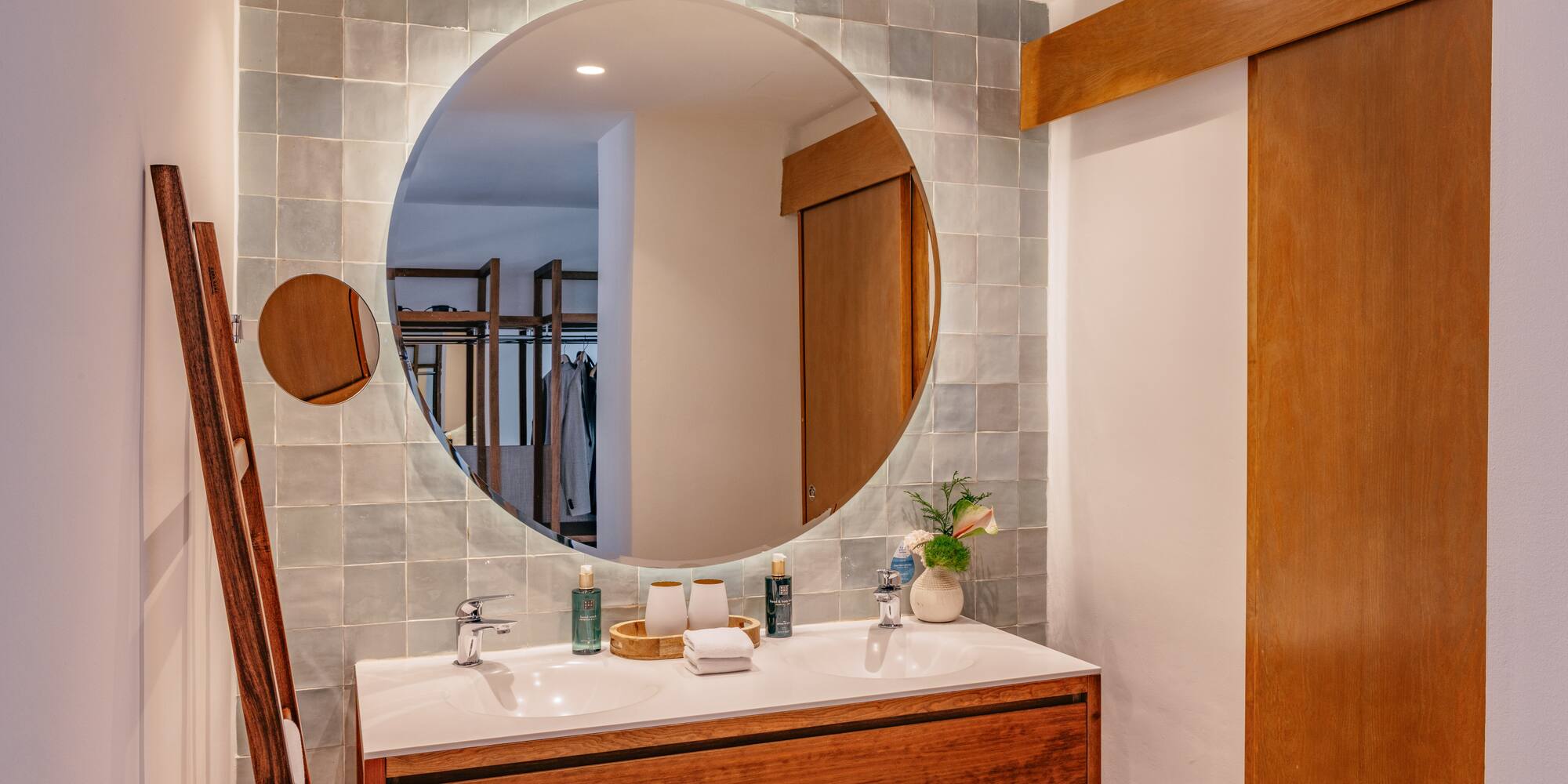 Double sink bathroom vanity with round backlit mirror and grey tiled wall.