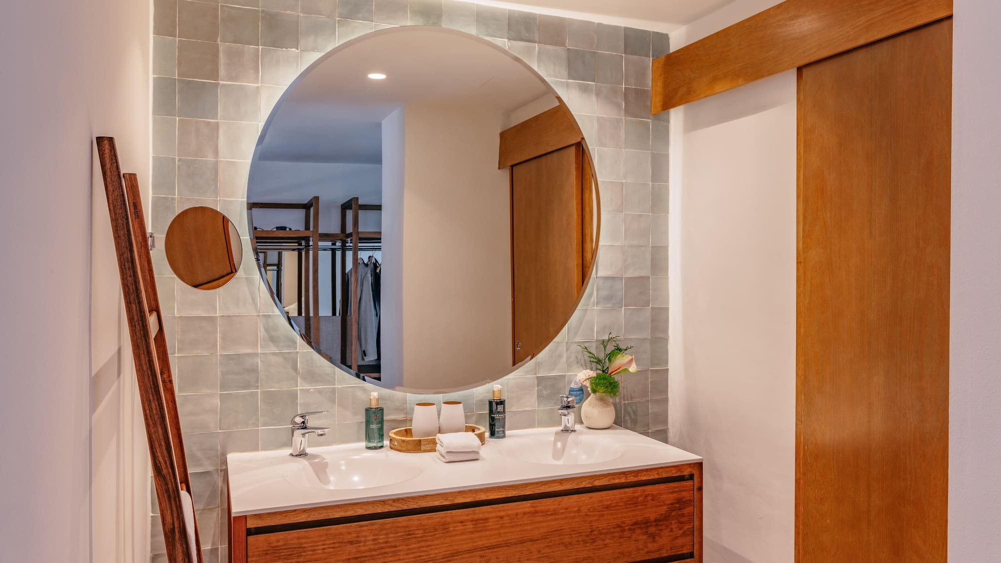 Double sink bathroom vanity with round backlit mirror and grey tiled wall.