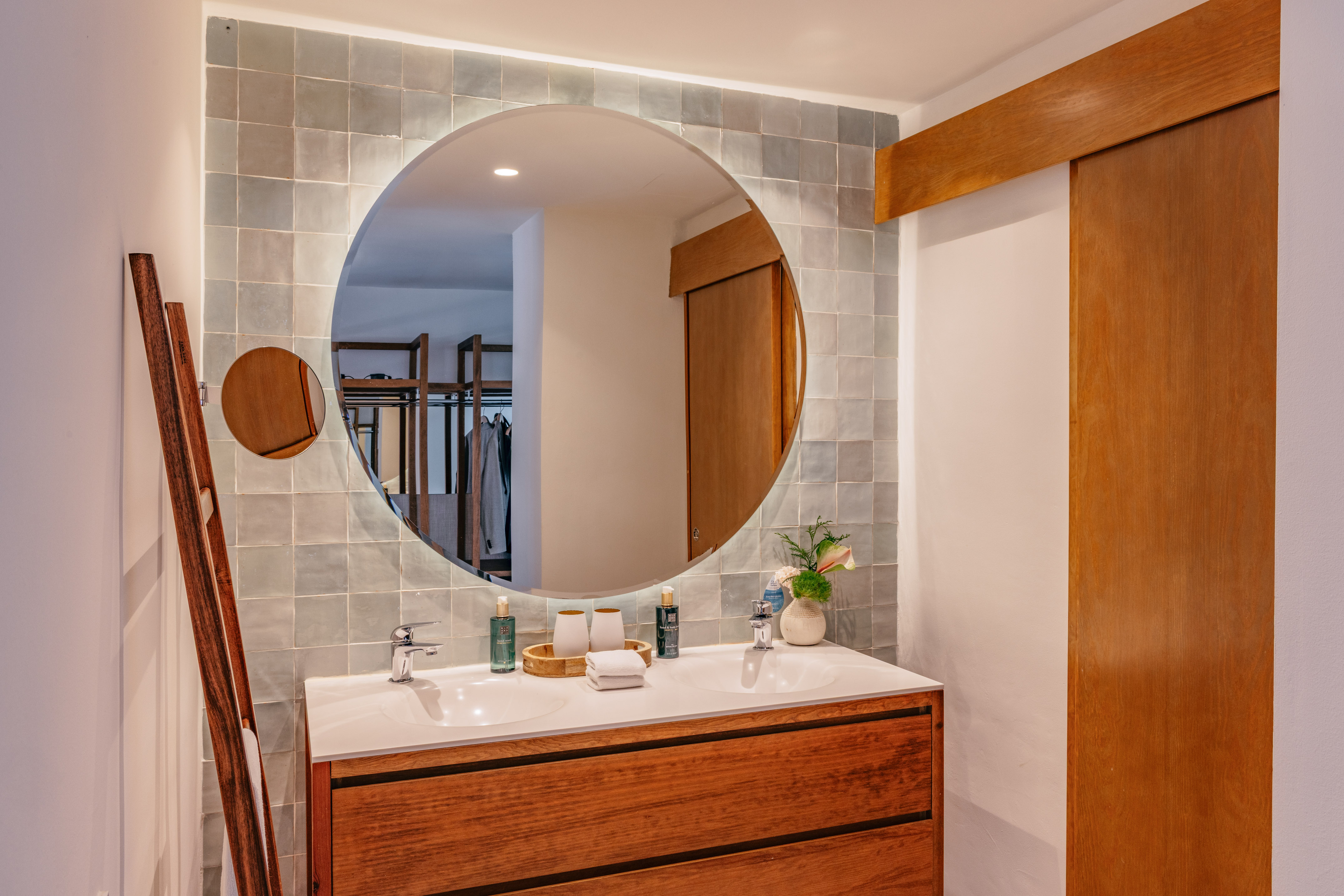 Double sink bathroom vanity with round backlit mirror and grey tiled wall.