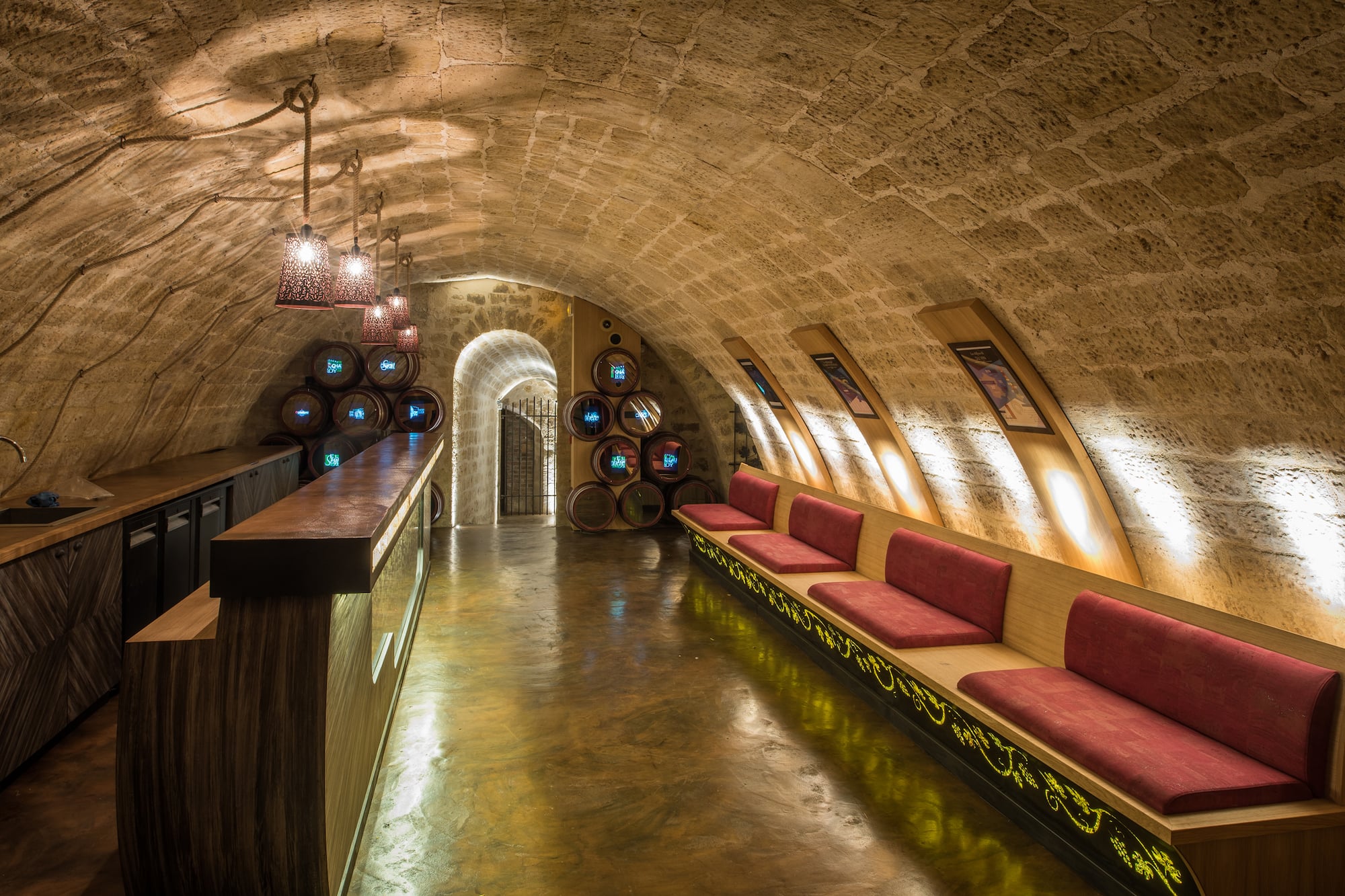 Bodega abovedada con bar, asientos rojos y barriles de vino iluminados.
