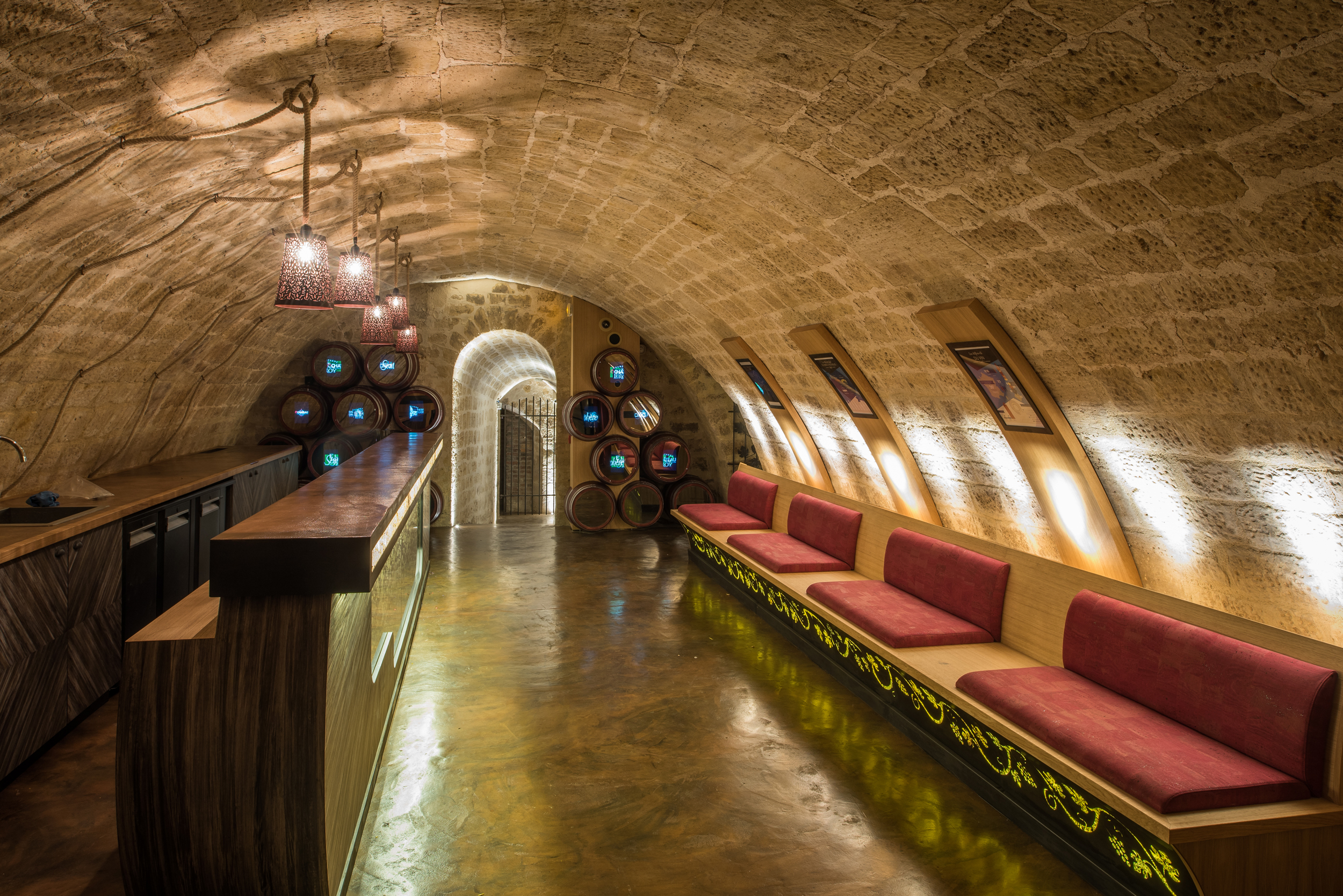 Bodega abovedada con bar, asientos rojos y barriles de vino iluminados.