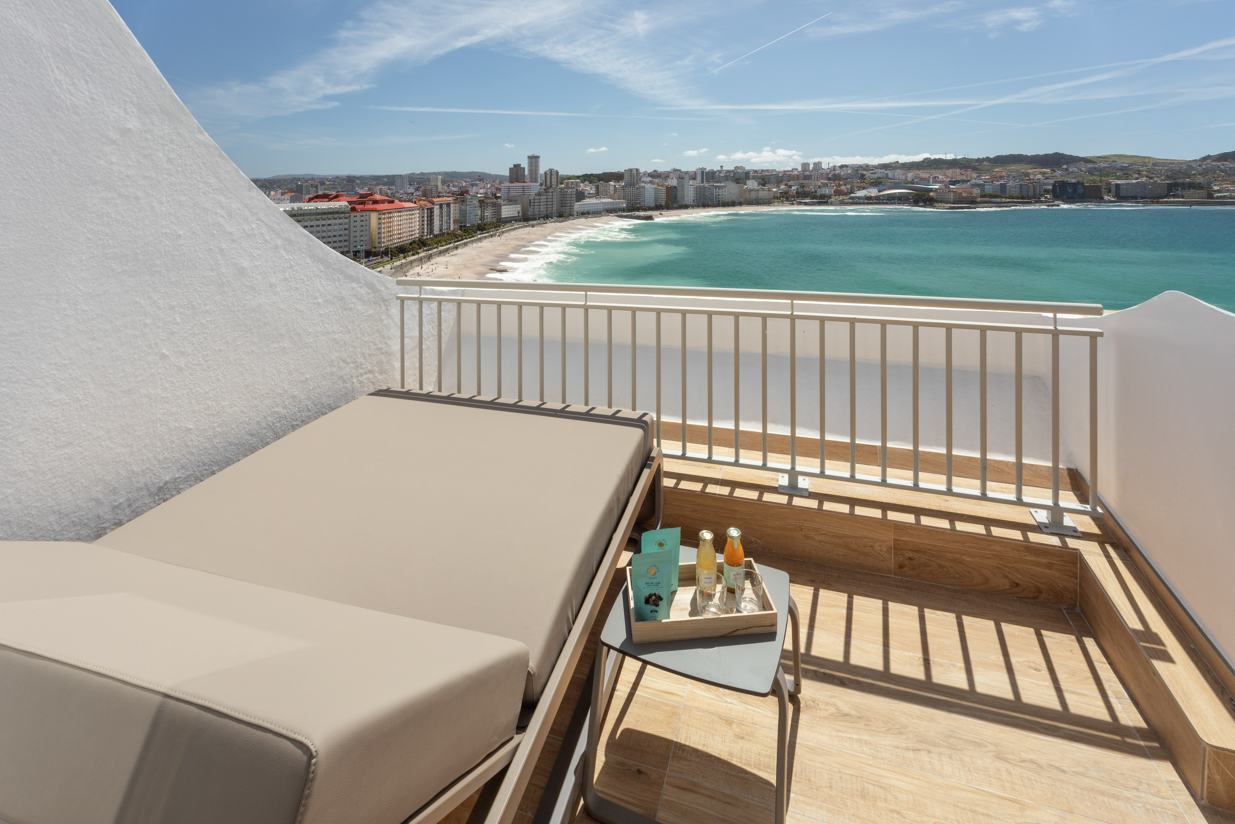 Sunny hotel balcony with lounge chair overlooking a sandy beach, ocean, and city.