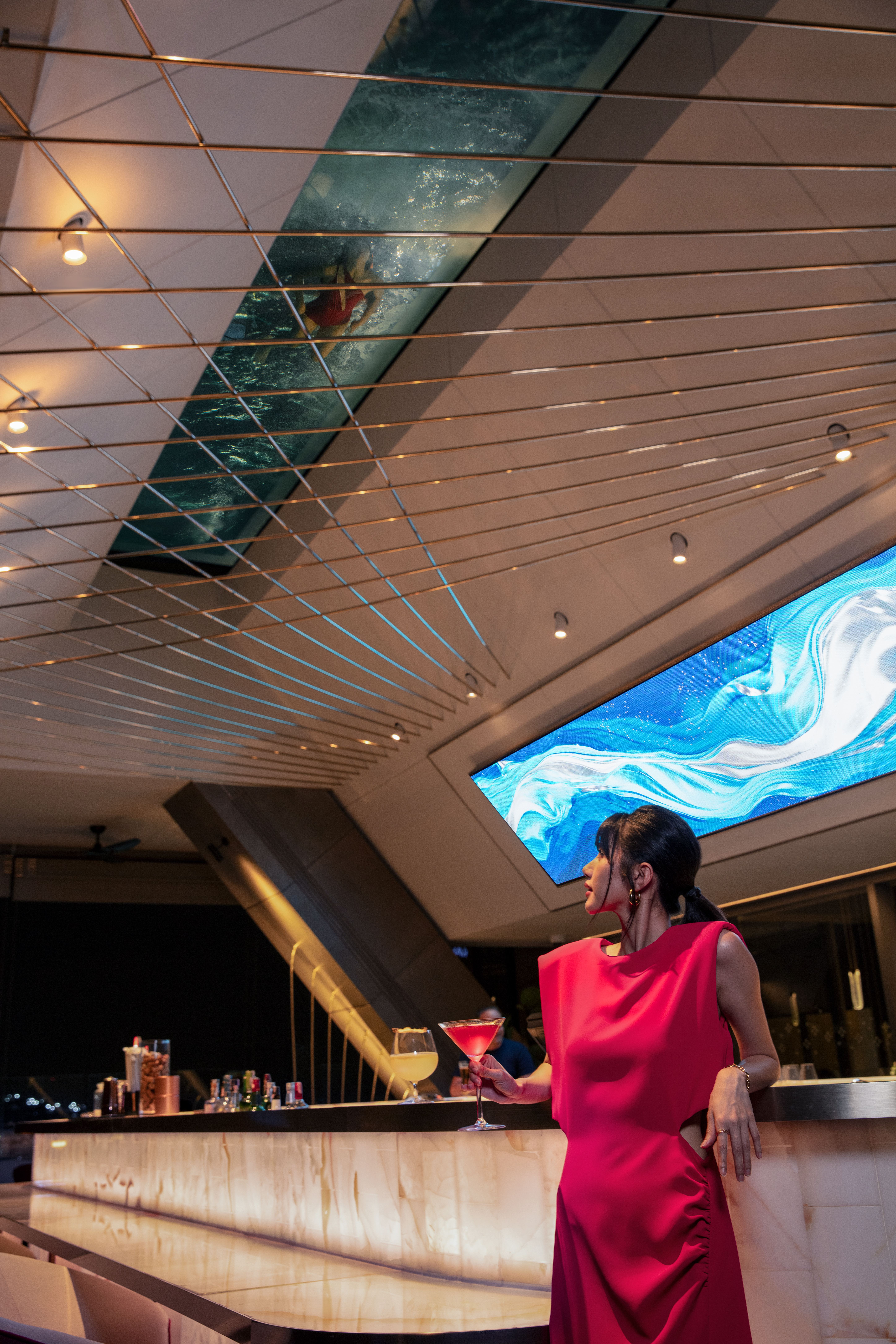 Woman in red dress at modern cocktail bar with overhead transparent pool.
