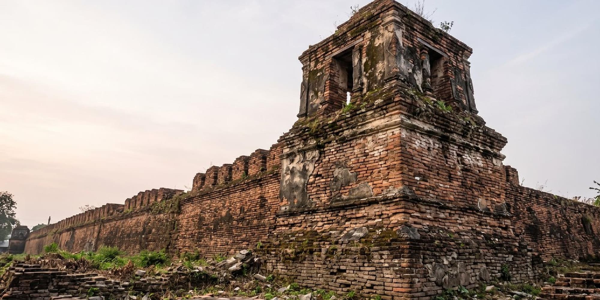 Muralla de ladrillo antigua y torreón en ruinas con vegetación.