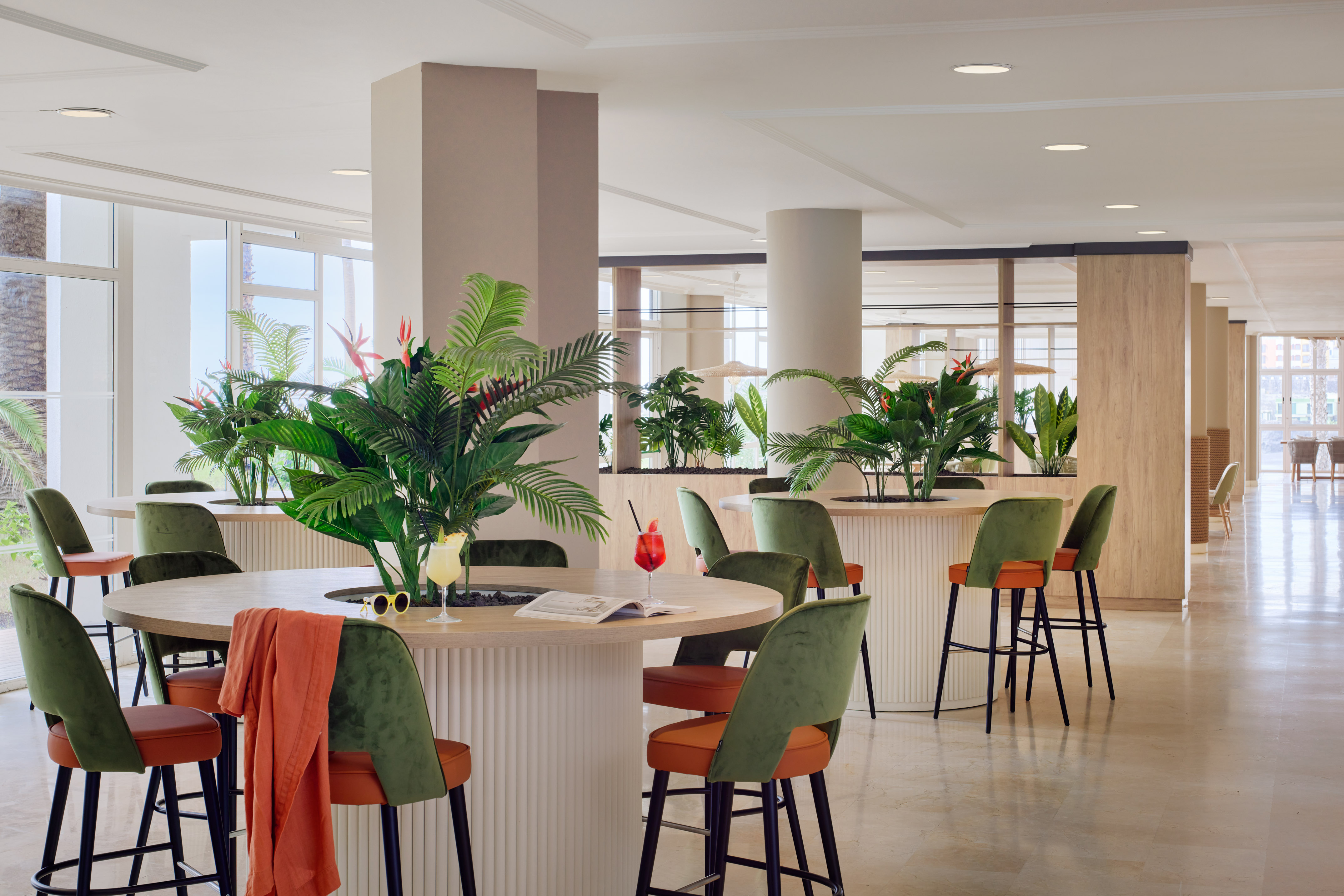 Modern hotel bar lounge with tropical plants and green-orange stools.