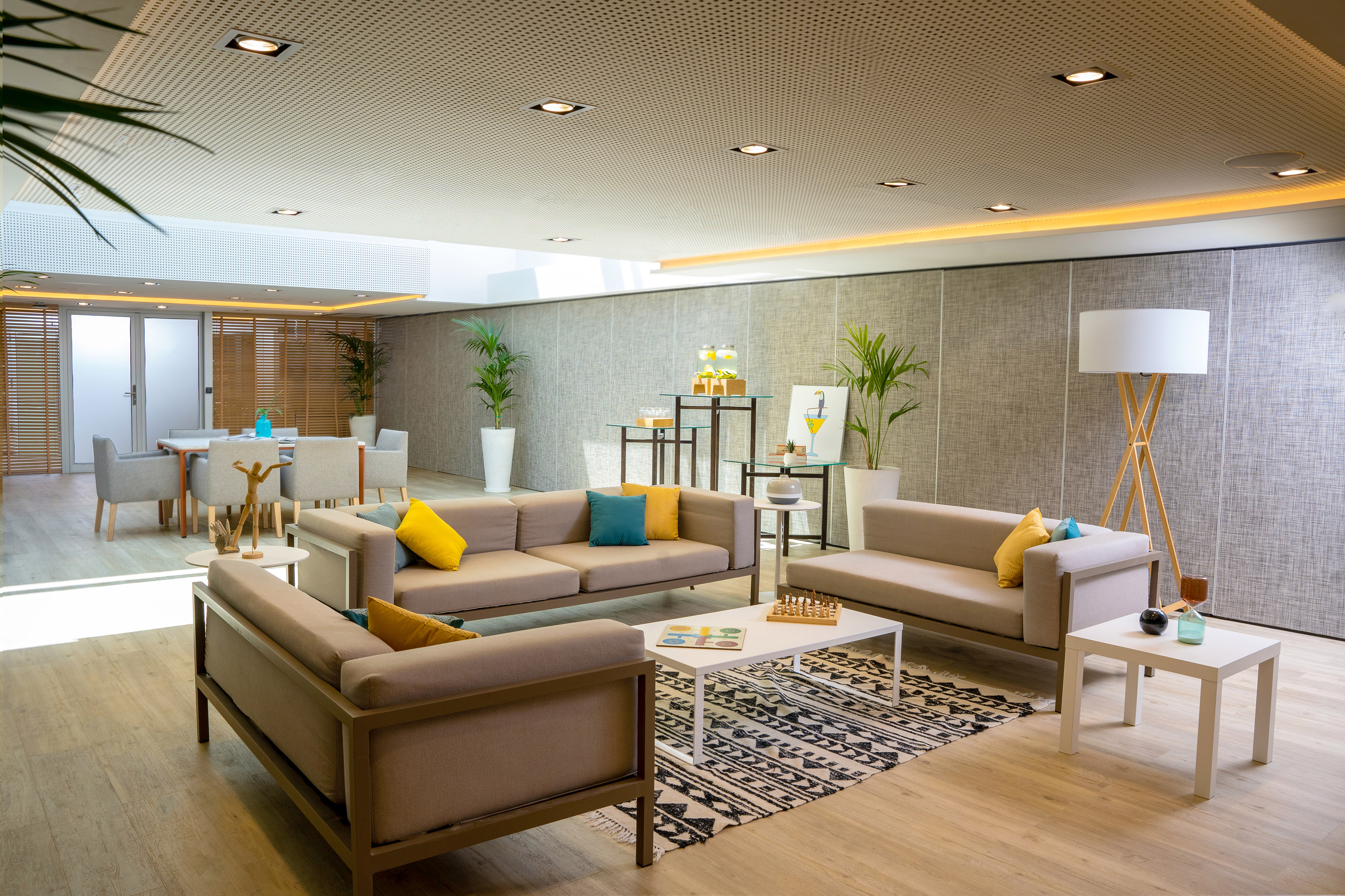 Contemporary lounge with beige sofas, colorful pillows, dining area, plants.