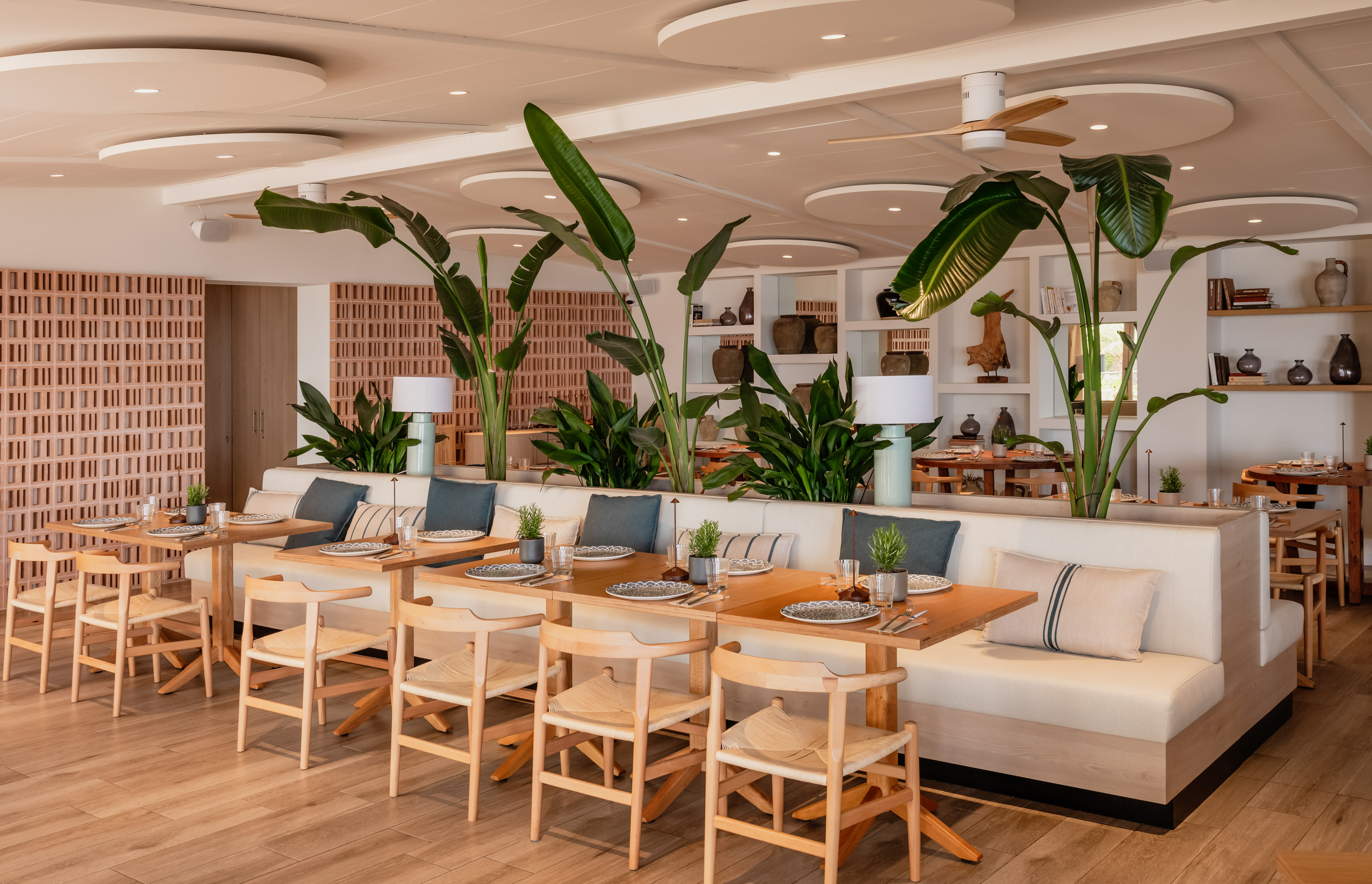 Modern restaurant dining room with wood tables, banquettes, and tropical plants.