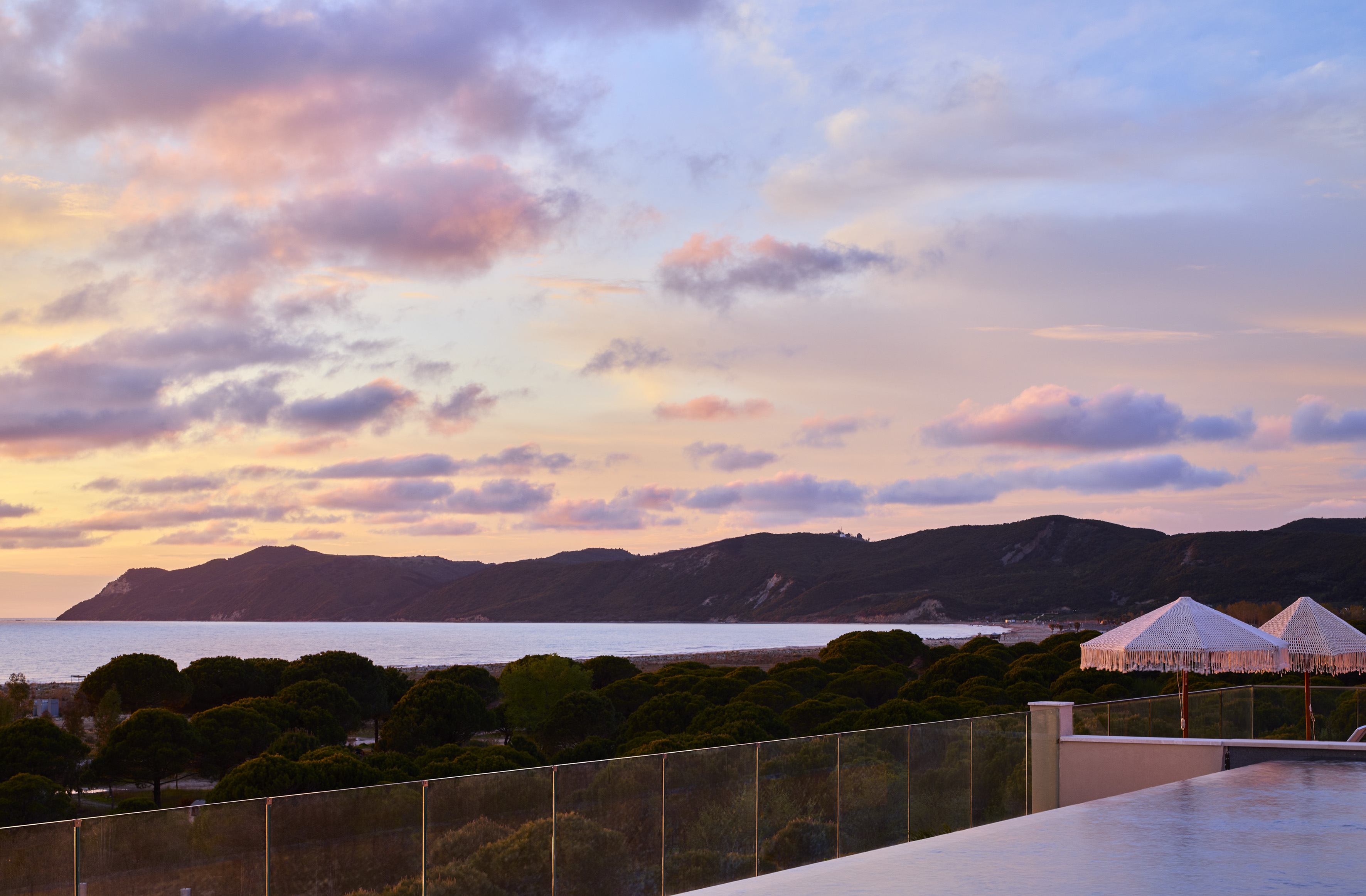 Vibrant sunset over coastal mountains, ocean, and infinity pool with umbrellas.
