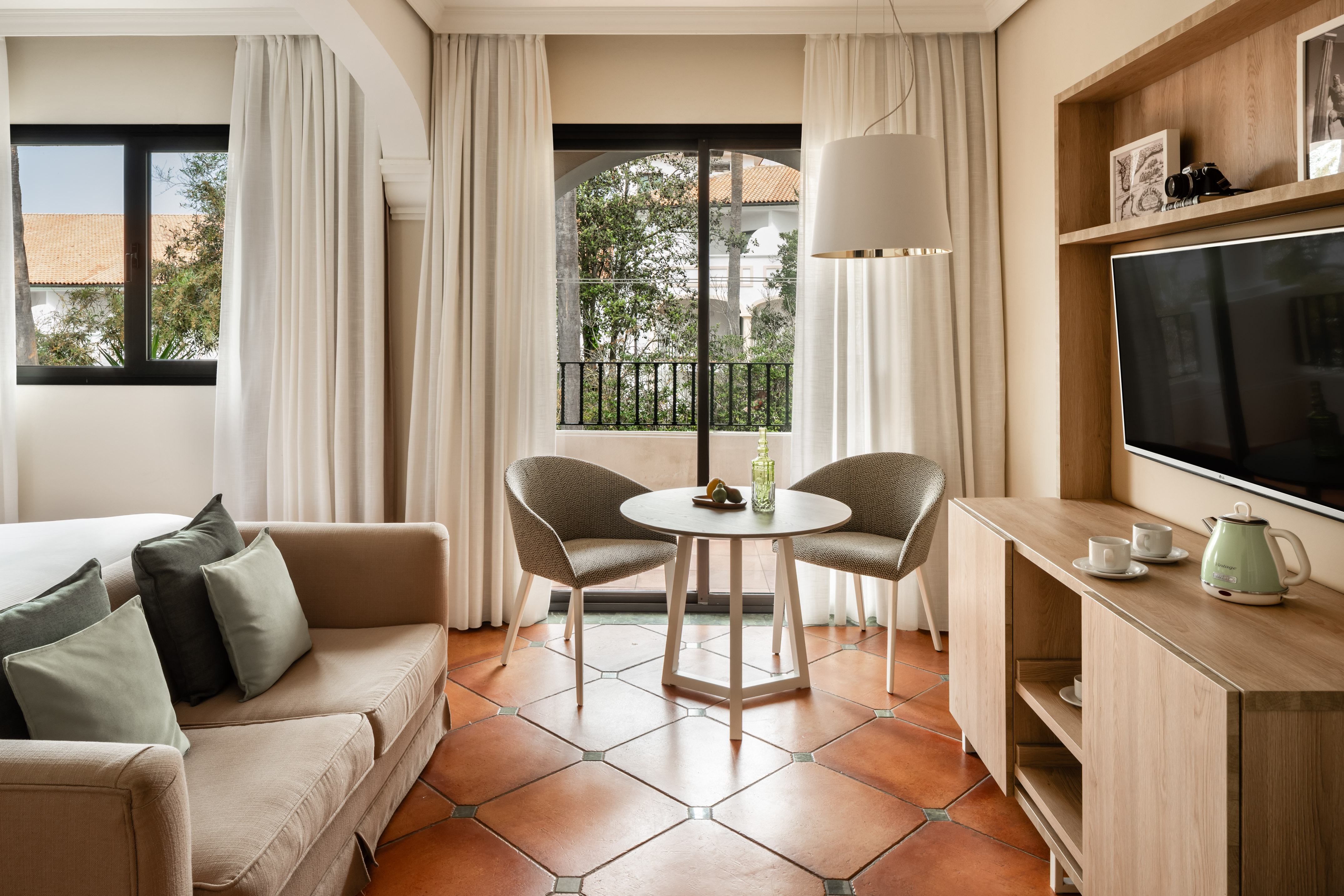 Modern hotel living area with balcony, sofa, TV, and terracotta floor.